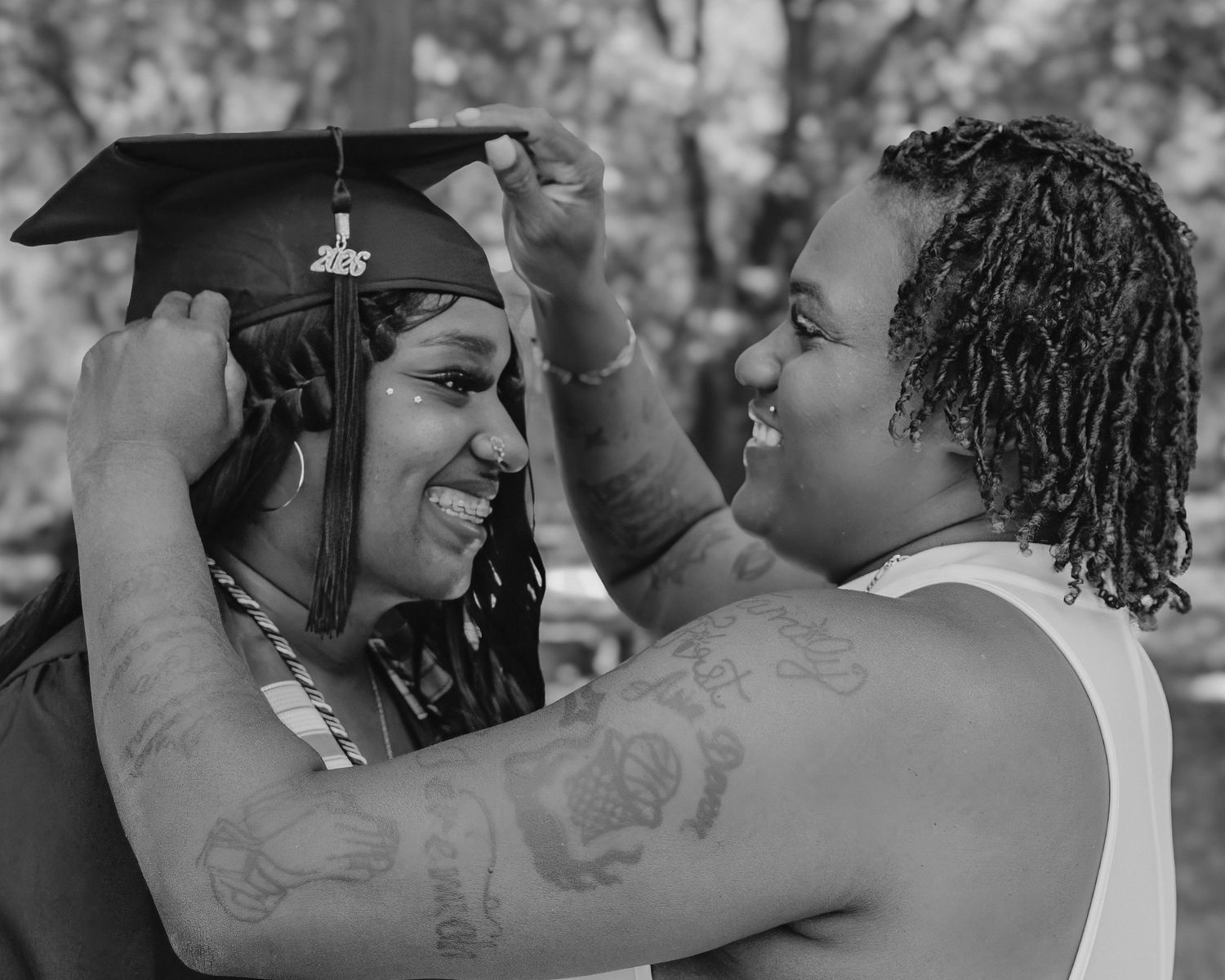 A graduate smiles as someone adjusts their cap during a black and white outdoor graduation celebration.