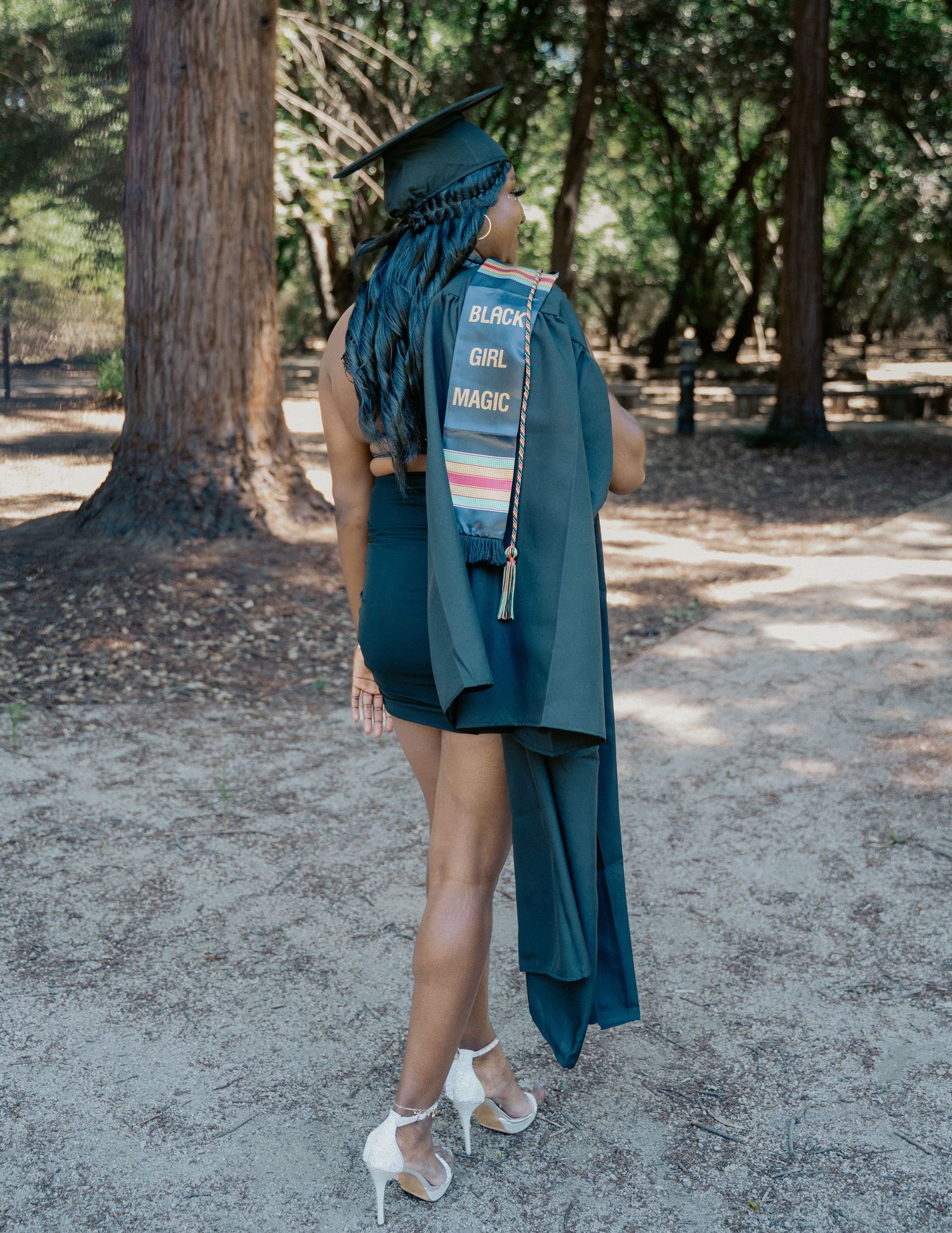 Graduate in teal cap and gown with Black Girl Magic stole poses outdoors among trees.