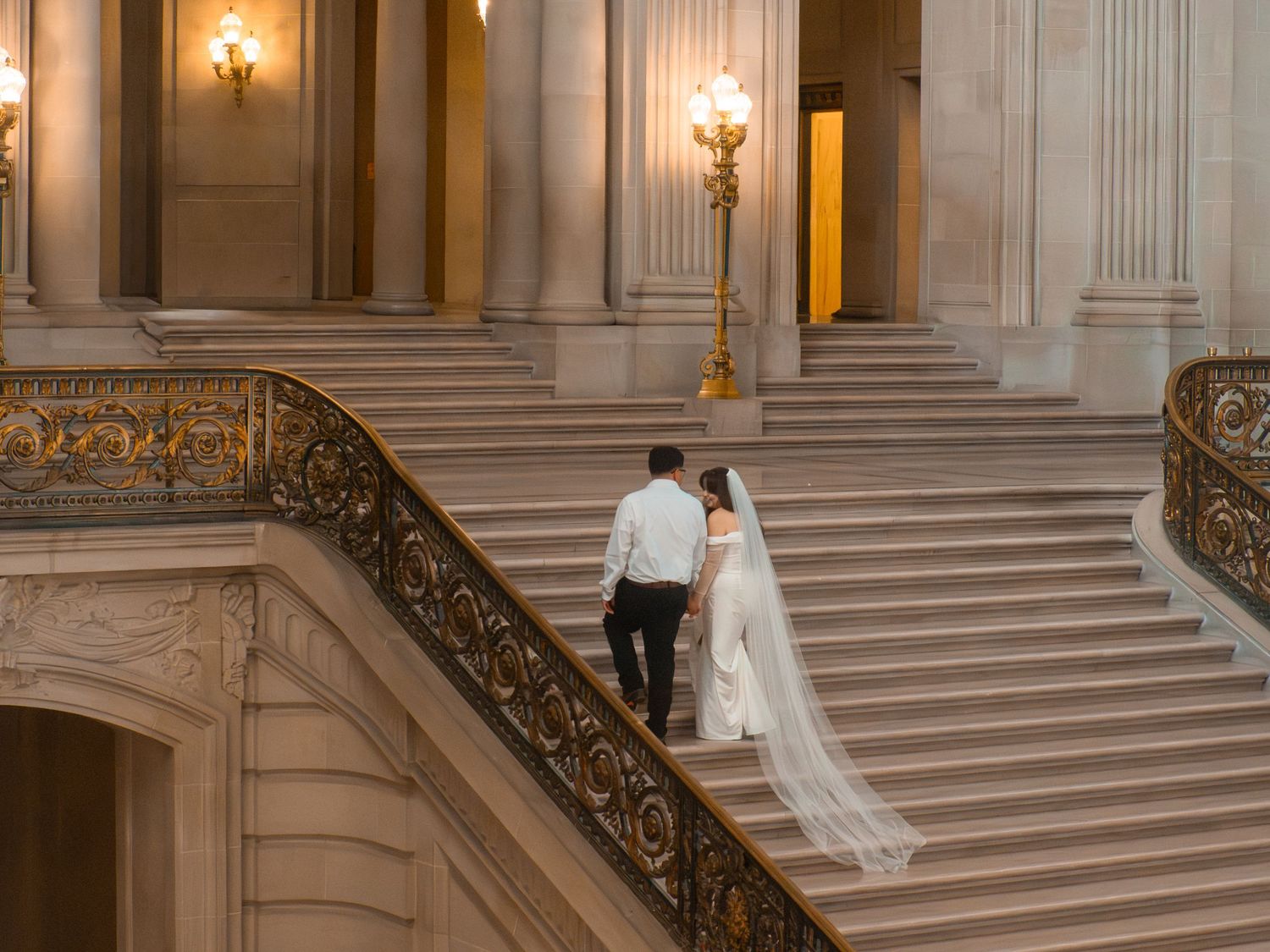 Bride and groom ascending grand staircase at San Francisco City Hall in elegant wedding attire.