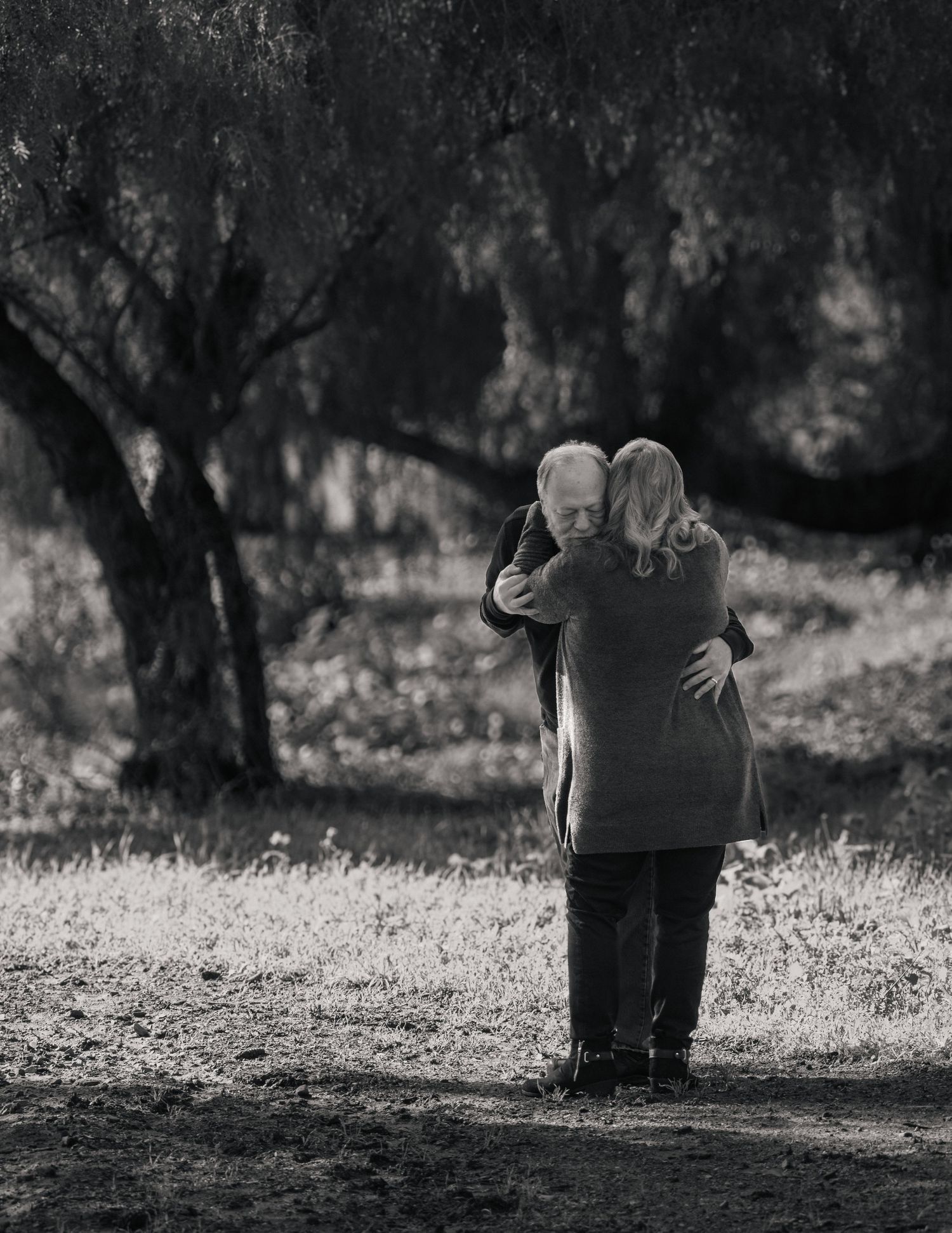 Black and white photo of an elderly couple embracing tenderly in a sunlit park surrounded by trees. Couples photography by Jolie & Rae Photo