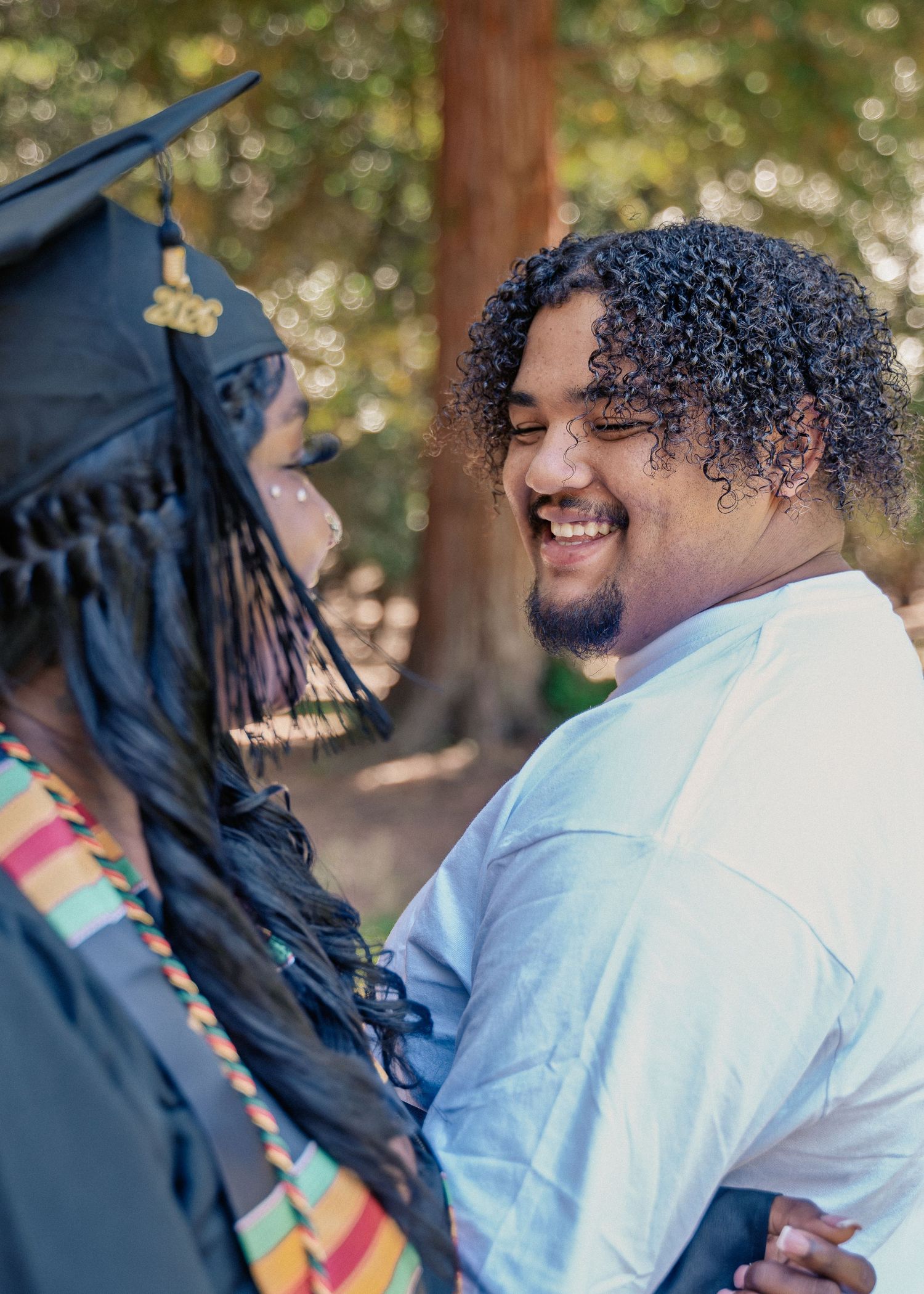Smiling young man embraces college graduate in cap and gown outdoors during graduation celebration.