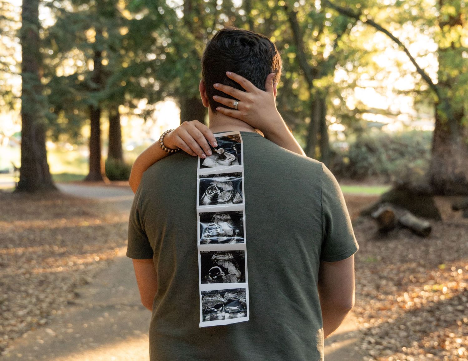 Couple embracing outdoors holding ultrasound photos, pregnancy announcement in a scenic park setting.