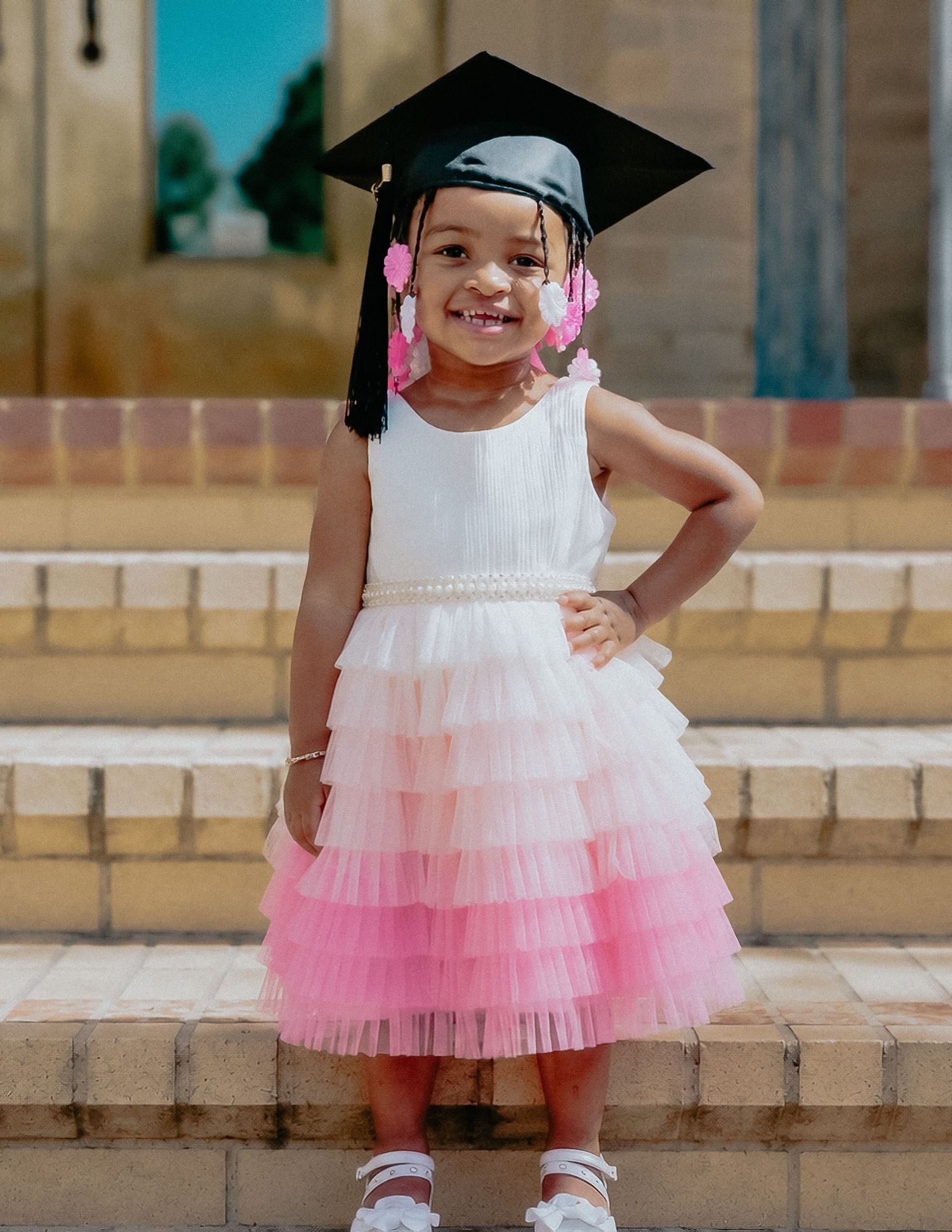 Smiling toddler girl in white pink tiered tulle dress and graduation cap posing on stone steps.