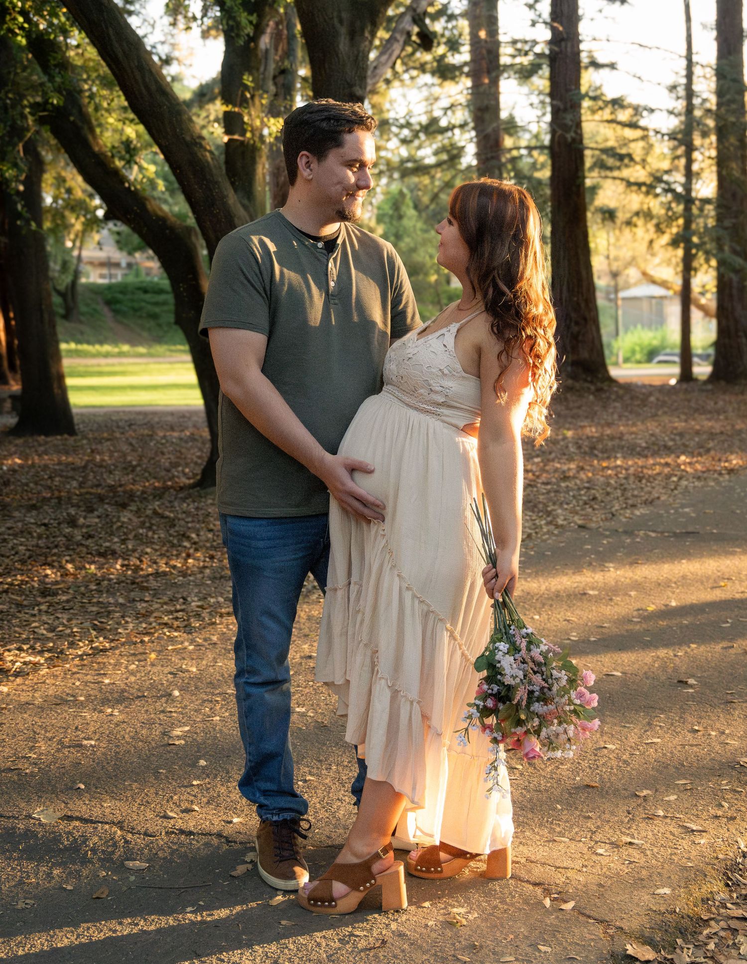 A couple walks hand in hand through a sunlit park with autumn leaves scattered on the ground during golden hour maternity portrait. Lodi, CA