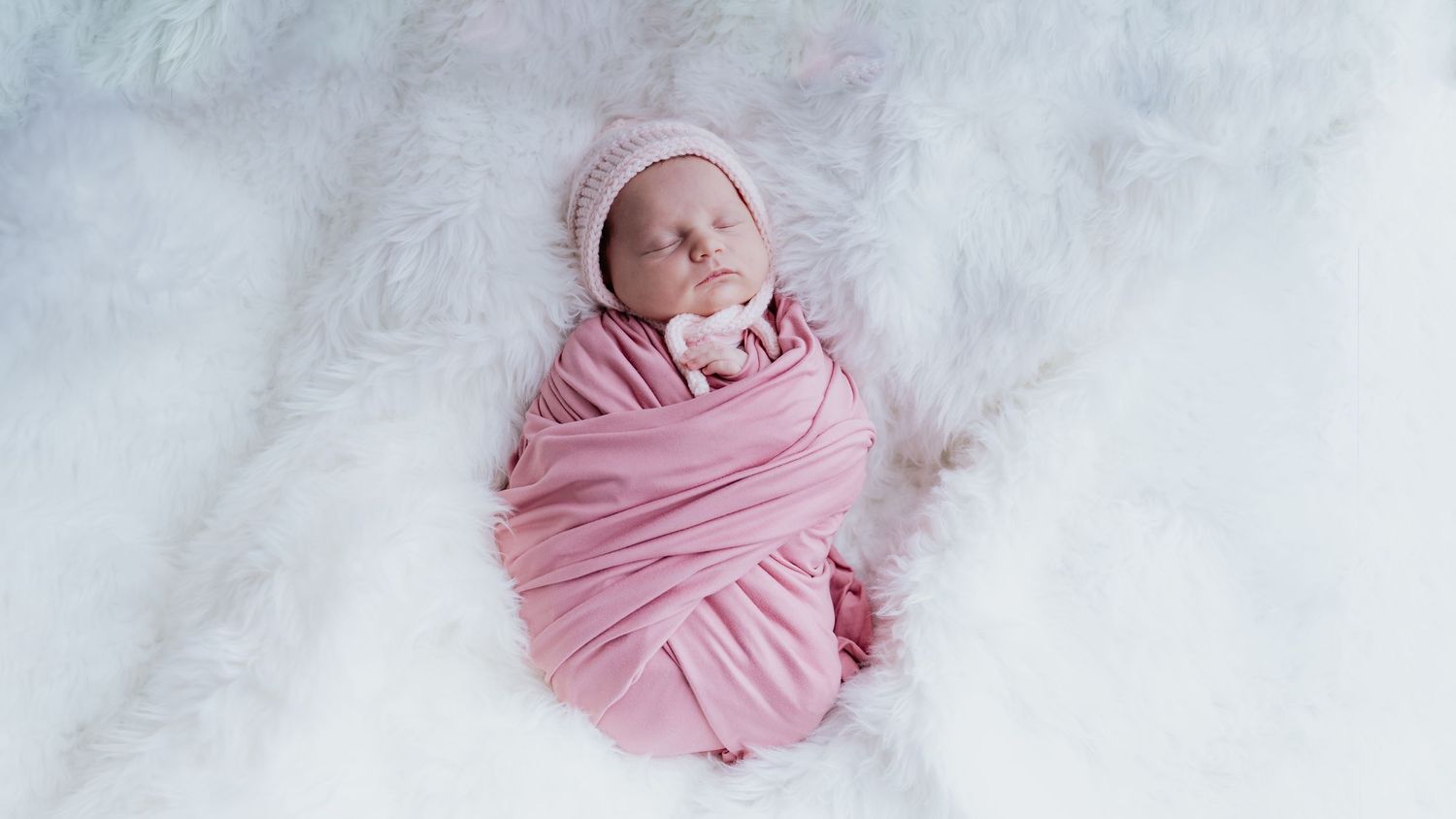 A newborn baby swaddled in a soft pink wrap and matching hat, resting peacefully on a white fur blanket. Newborn photography near Folsom, CA