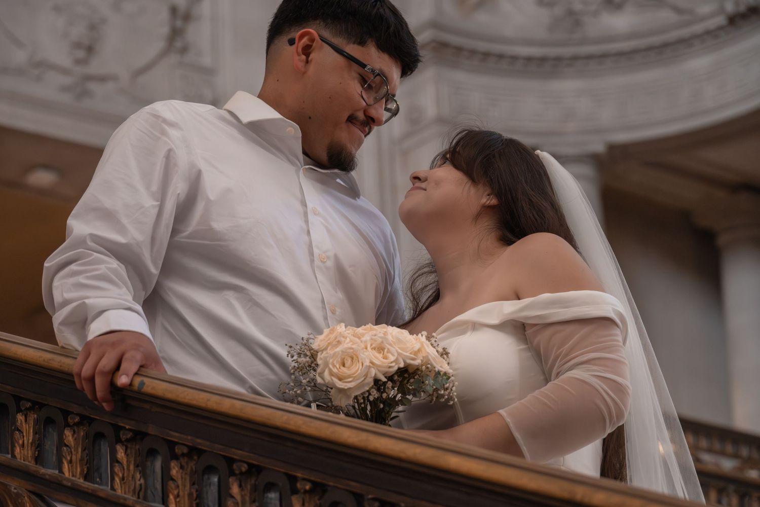 Newlywed couple in wedding attire sharing a romantic moment on a balcony, bride holding white roses bouquet.