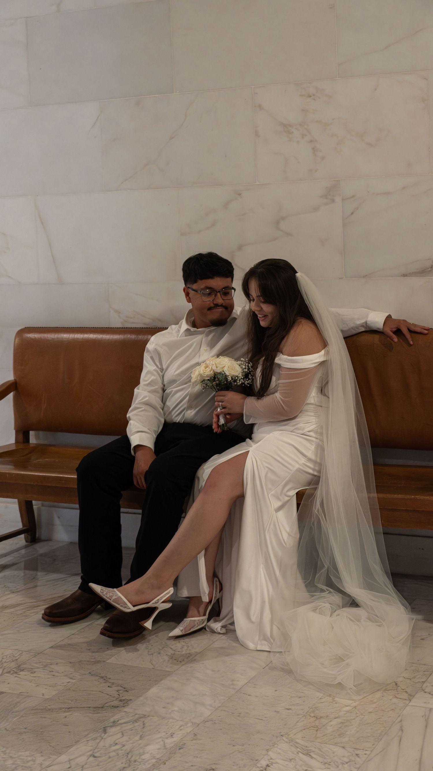 A bride in white gown and veil sits with groom on wooden bench, sharing a tender moment in marble hall.