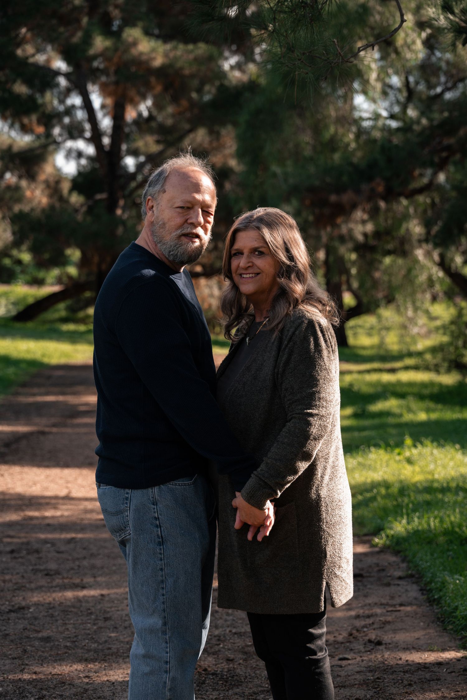 Middle-aged couple holding hands on a park path surrounded by pine trees on a sunny day.
