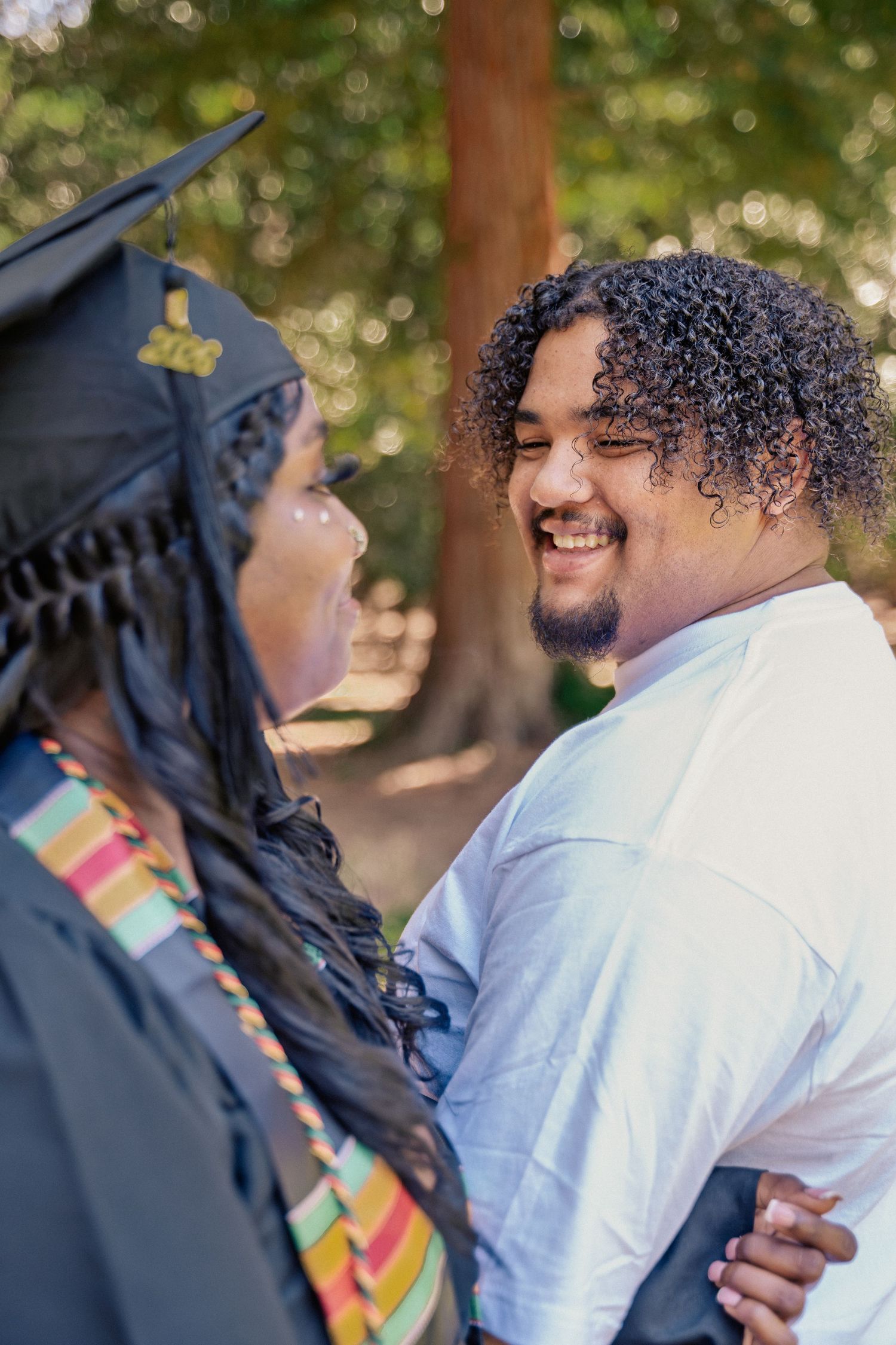 A smiling man with curly hair embraces a graduate wearing a cap and kente stole outdoors.