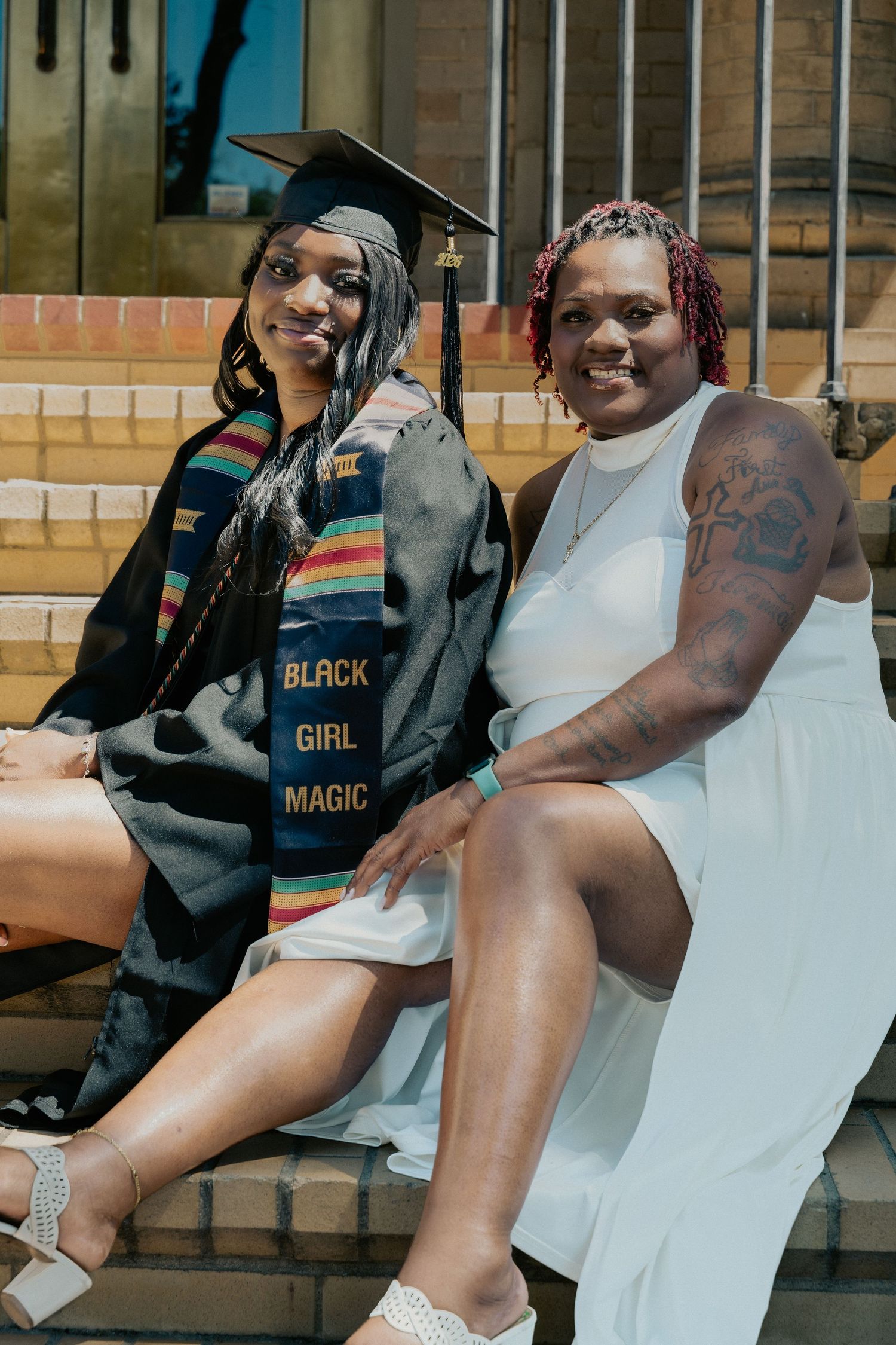 Two women sitting on steps, one wearing graduation cap, gown, and Black Girl Magic stole, celebrating academic achievement.