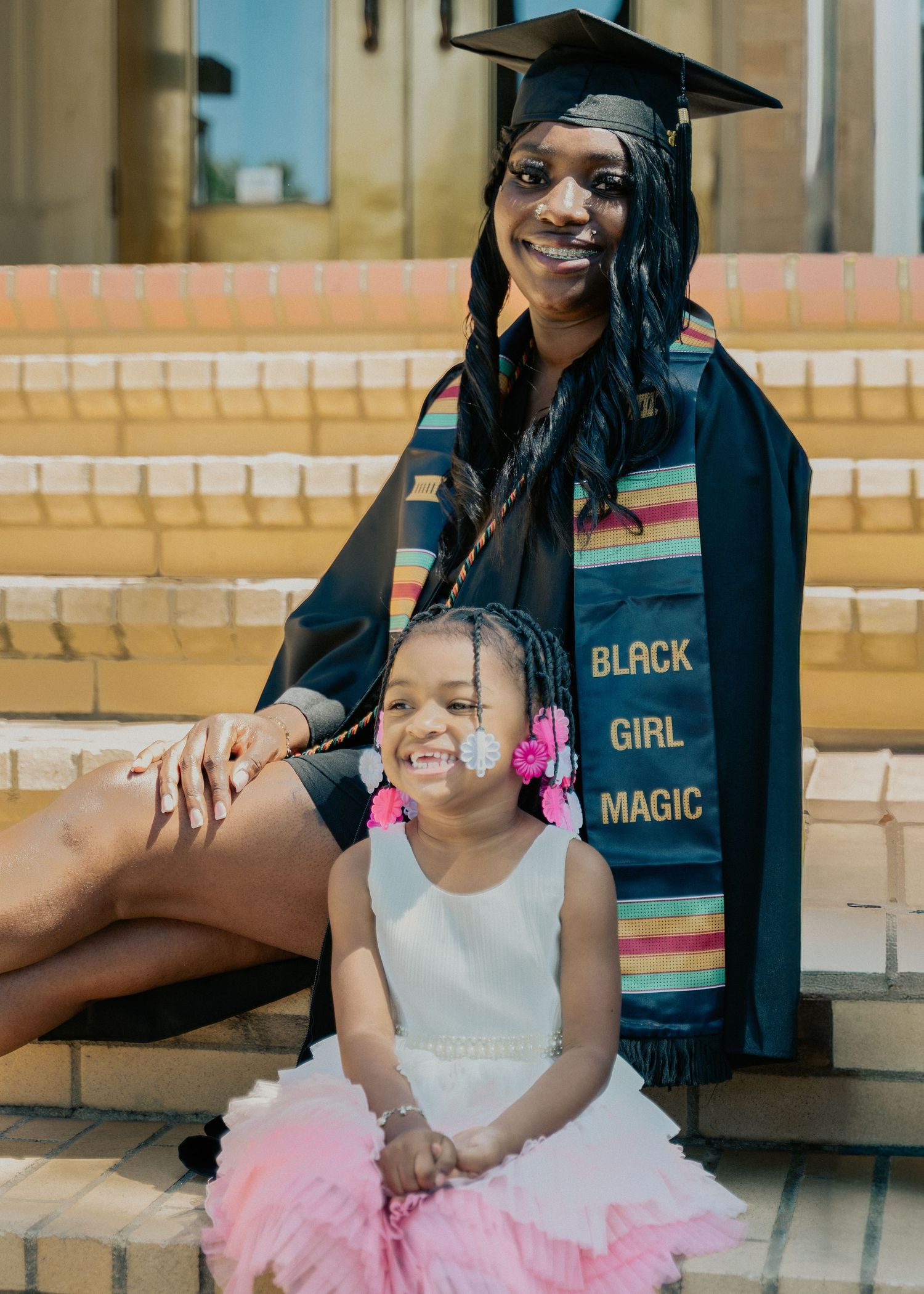 Graduate in cap, gown, and Black Girl Magic stole poses with young girl in pink tutu on steps.