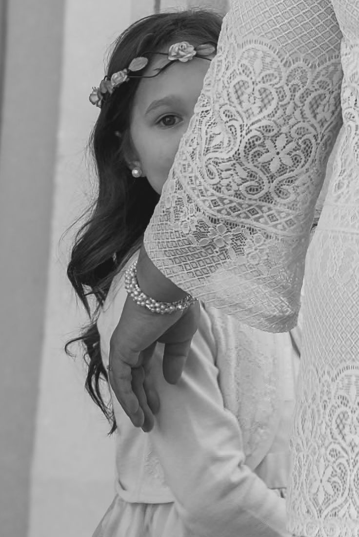 Black and white wedding photo of a young flower girl peeking from behind a bride’s lace sleeve, wearing a floral headband and pearl earrings