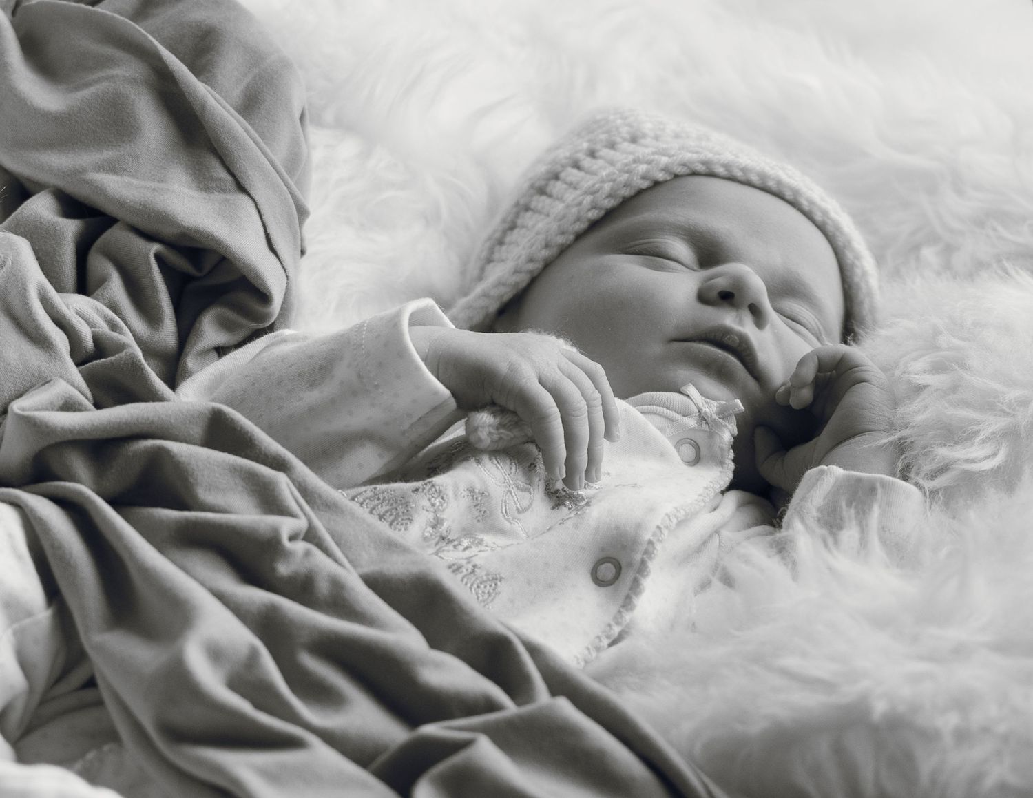 Peaceful newborn baby sleeping on fluffy white fur blanket wearing knit hat, black and white photography.