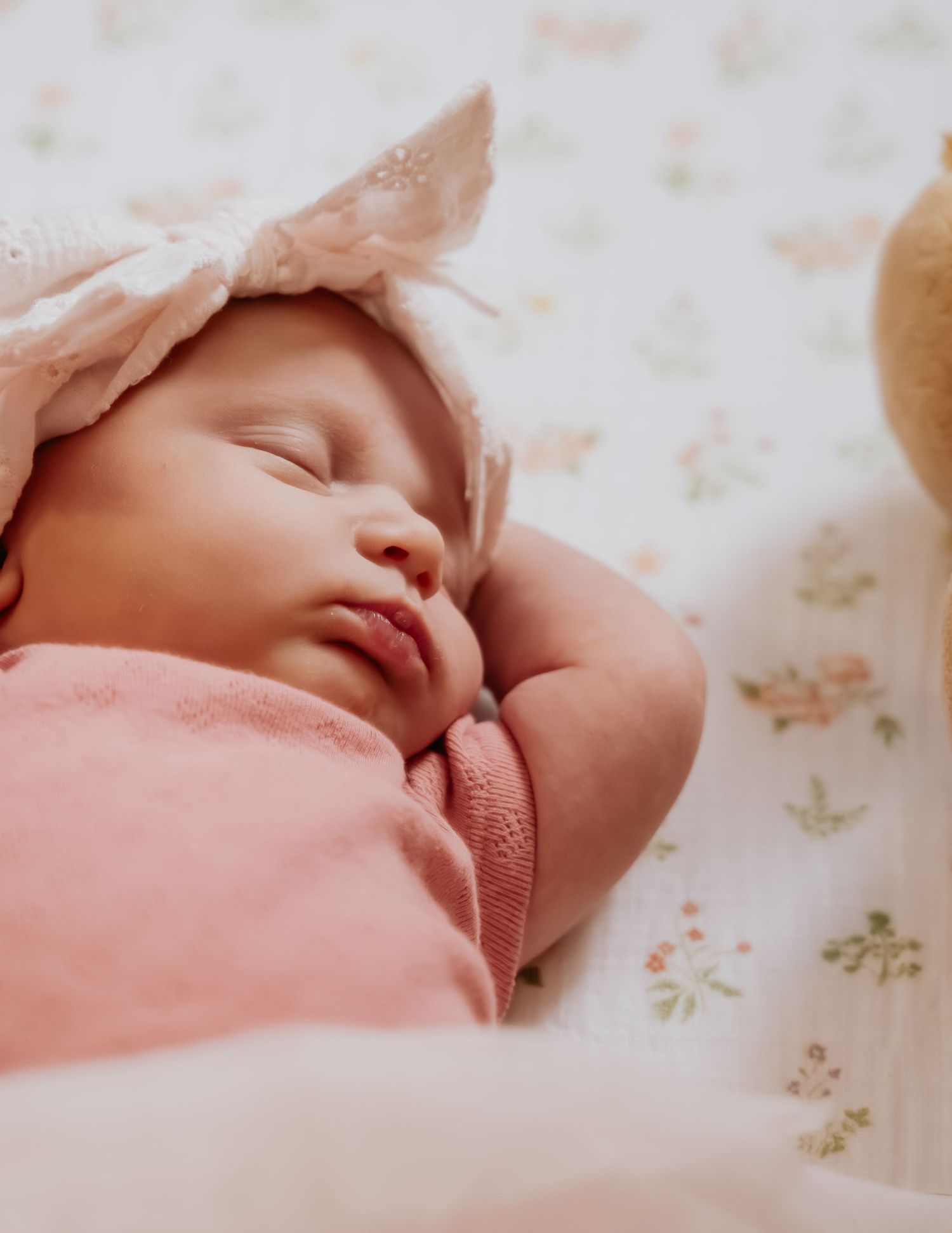 Peaceful newborn baby girl sleeping in pink outfit and bonnet on floral bedding.