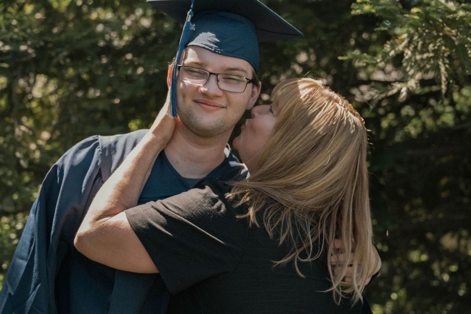 A proud graduate in cap and gown smiles as a woman kisses his cheek outdoors near green trees.