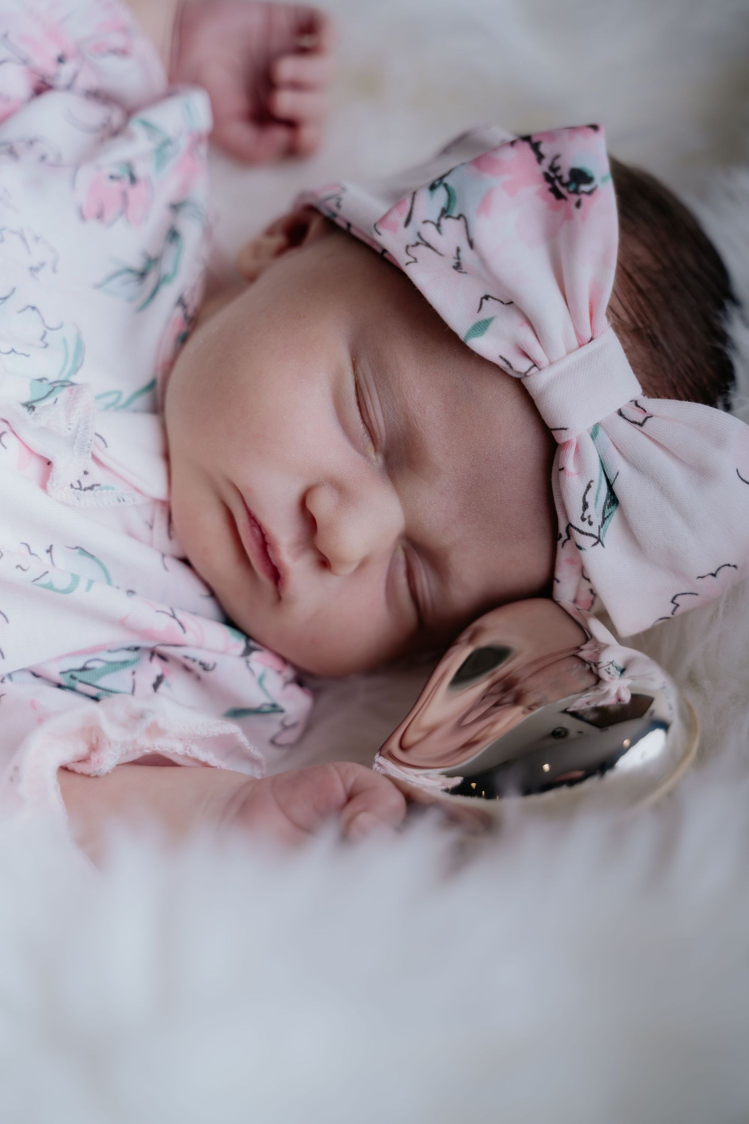 Sleeping newborn baby girl in pink floral outfit with matching bow headband, holding a silver pacifier.