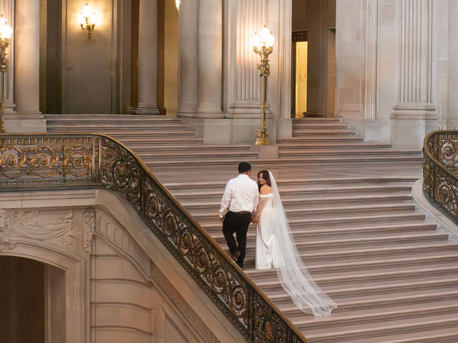A couple descends a grand marble staircase with ornate railings in a historic building.