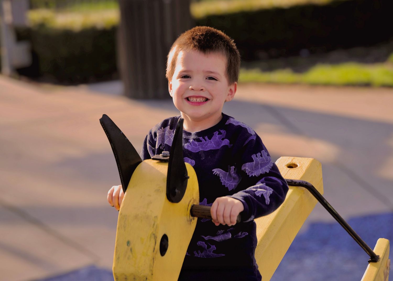 A child wearing a purple sweater rides a yellow rocking horse toy outdoors.