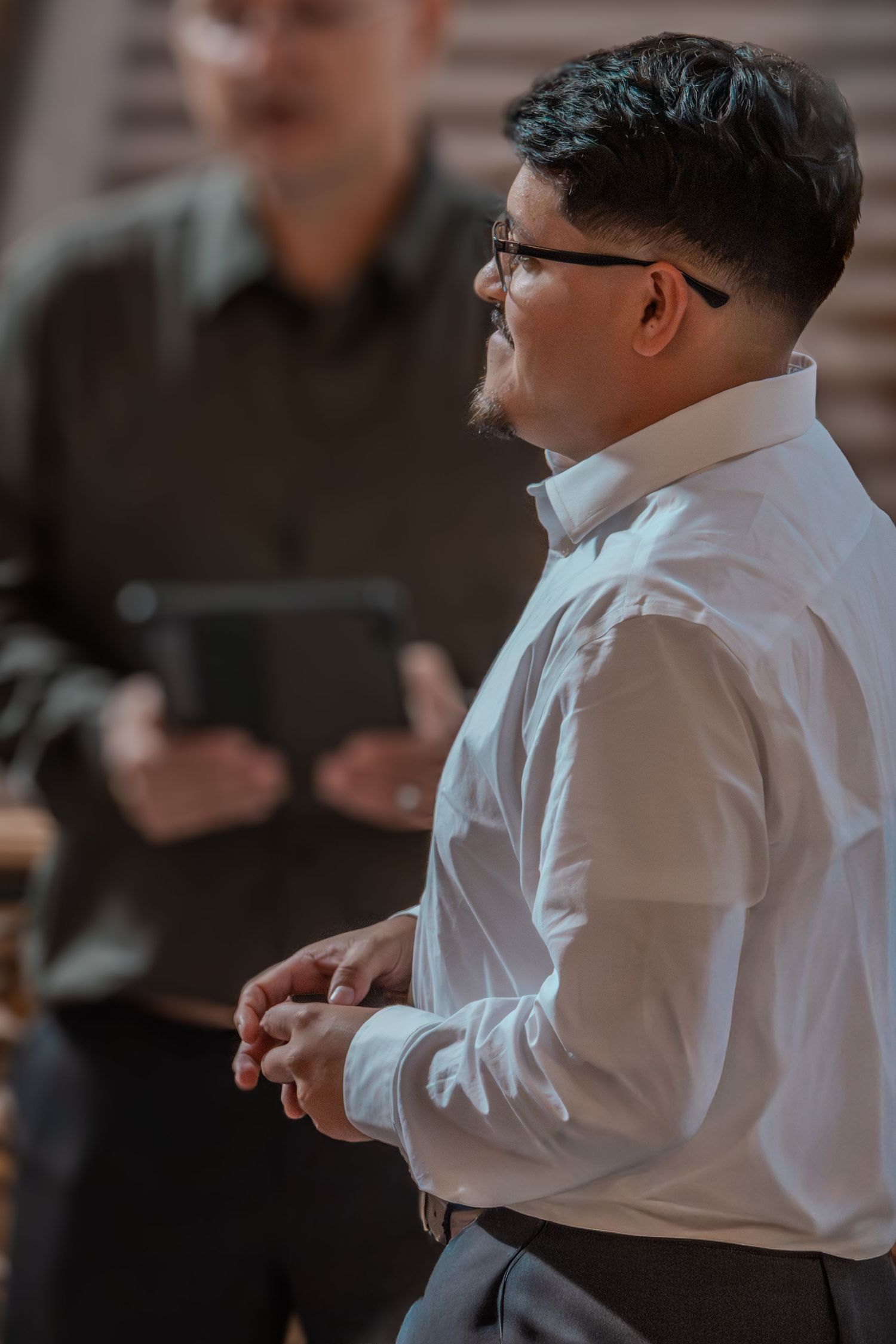 A person in a white dress shirt and glasses stands in conversation during an indoor event.