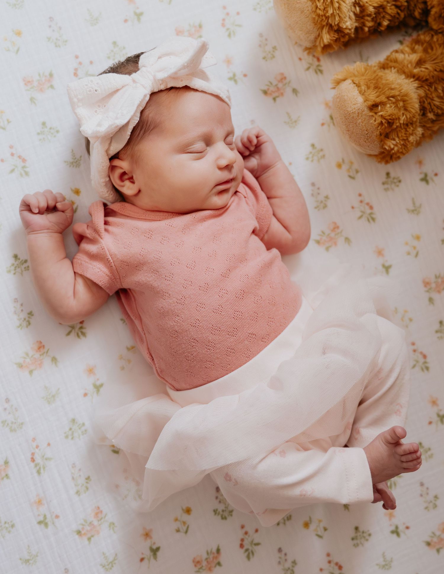 Sleeping newborn baby girl in pink outfit and bow headband lying on floral sheet with teddy bear during newborn photography session, Lodi CA
