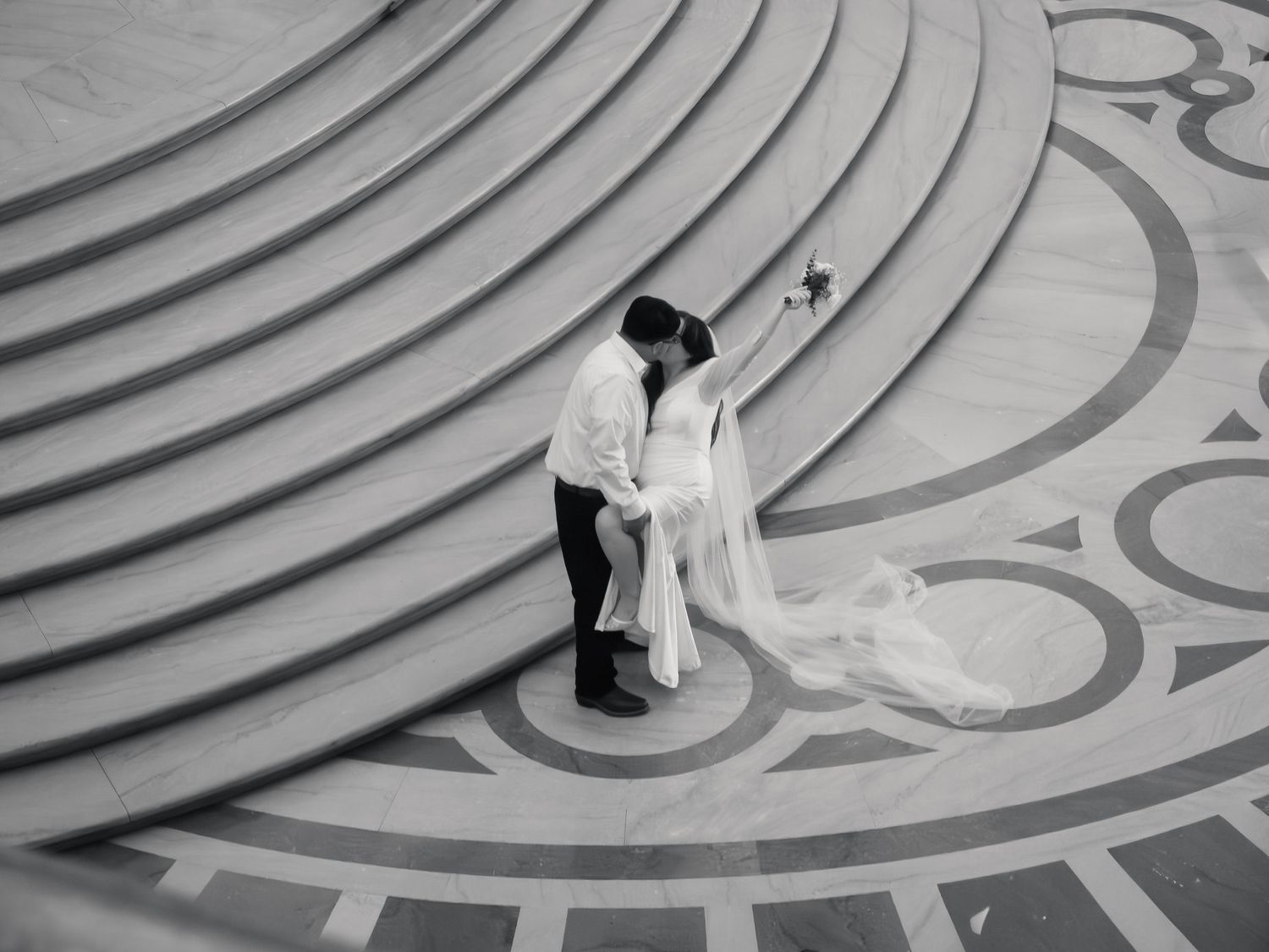 Black and white aerial photo of a bride and groom kissing on ornate circular marble steps at their wedding.