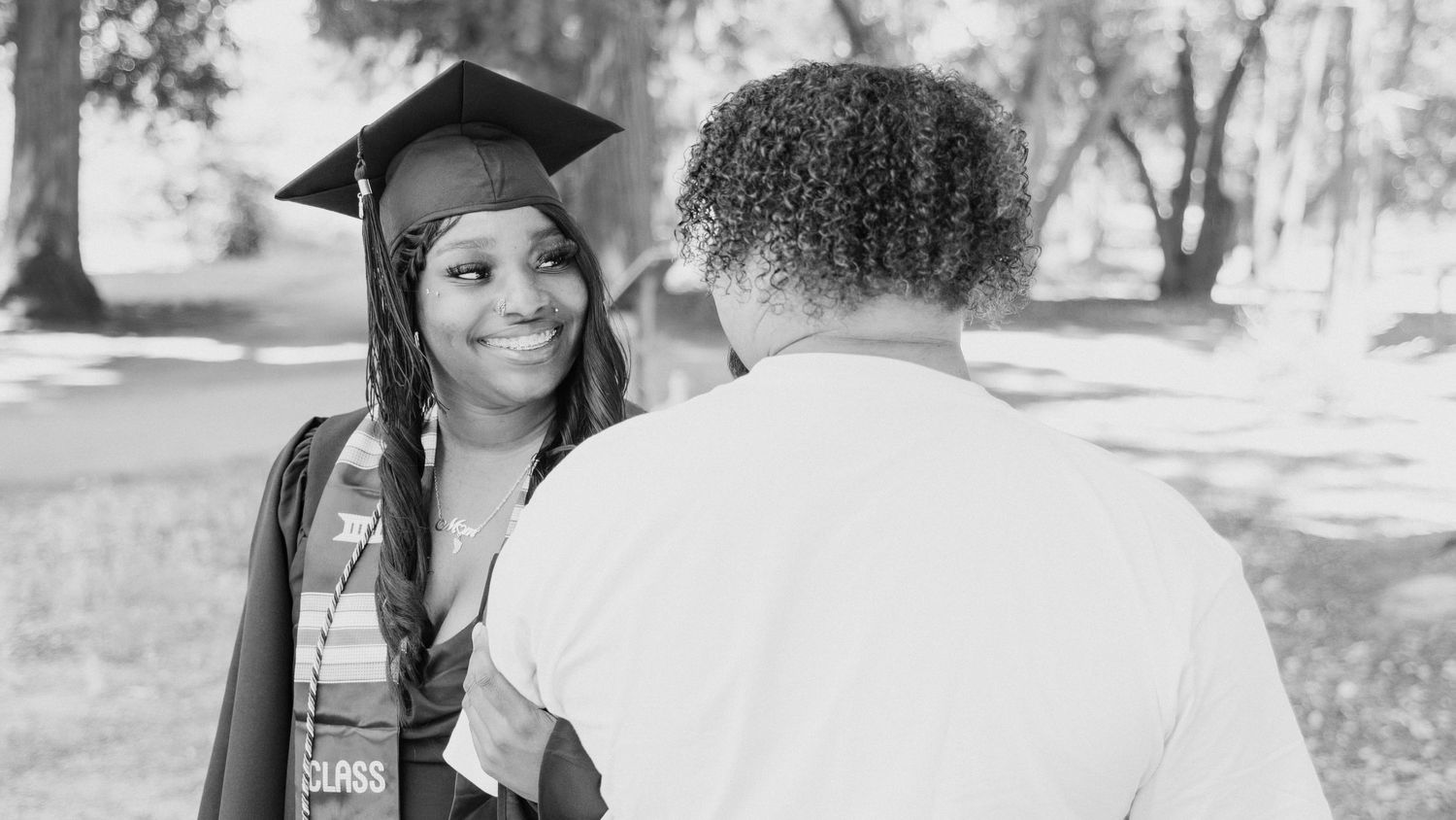 A smiling graduate in cap and gown shares a proud moment with a family member outdoors in a black and white photo.