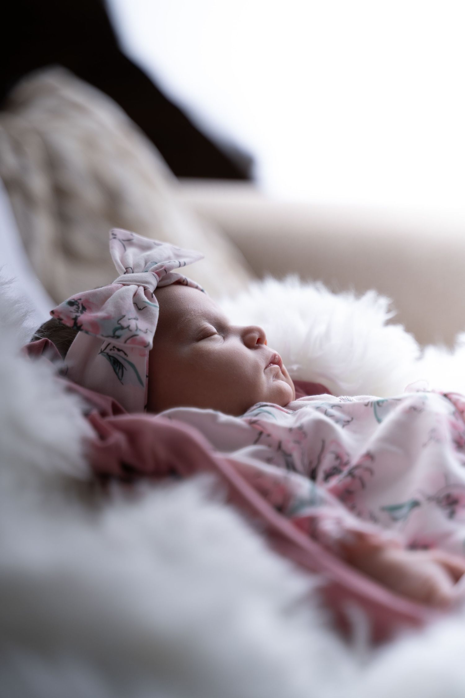 Sleeping newborn baby girl wrapped in pink floral swaddle blanket with matching bow headband on white fluffy surface. Roseville, CA