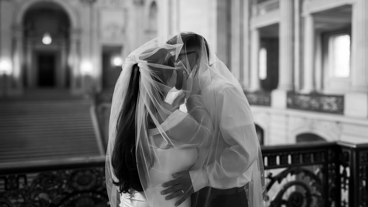 Black and white wedding photo of bride and groom kissing under veil in elegant grand hall.