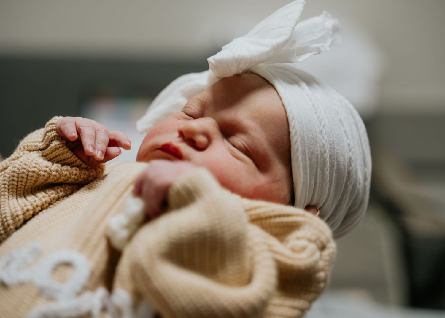 Peaceful newborn baby sleeping wrapped in cream blanket wearing white bow headband in hospital nursery.