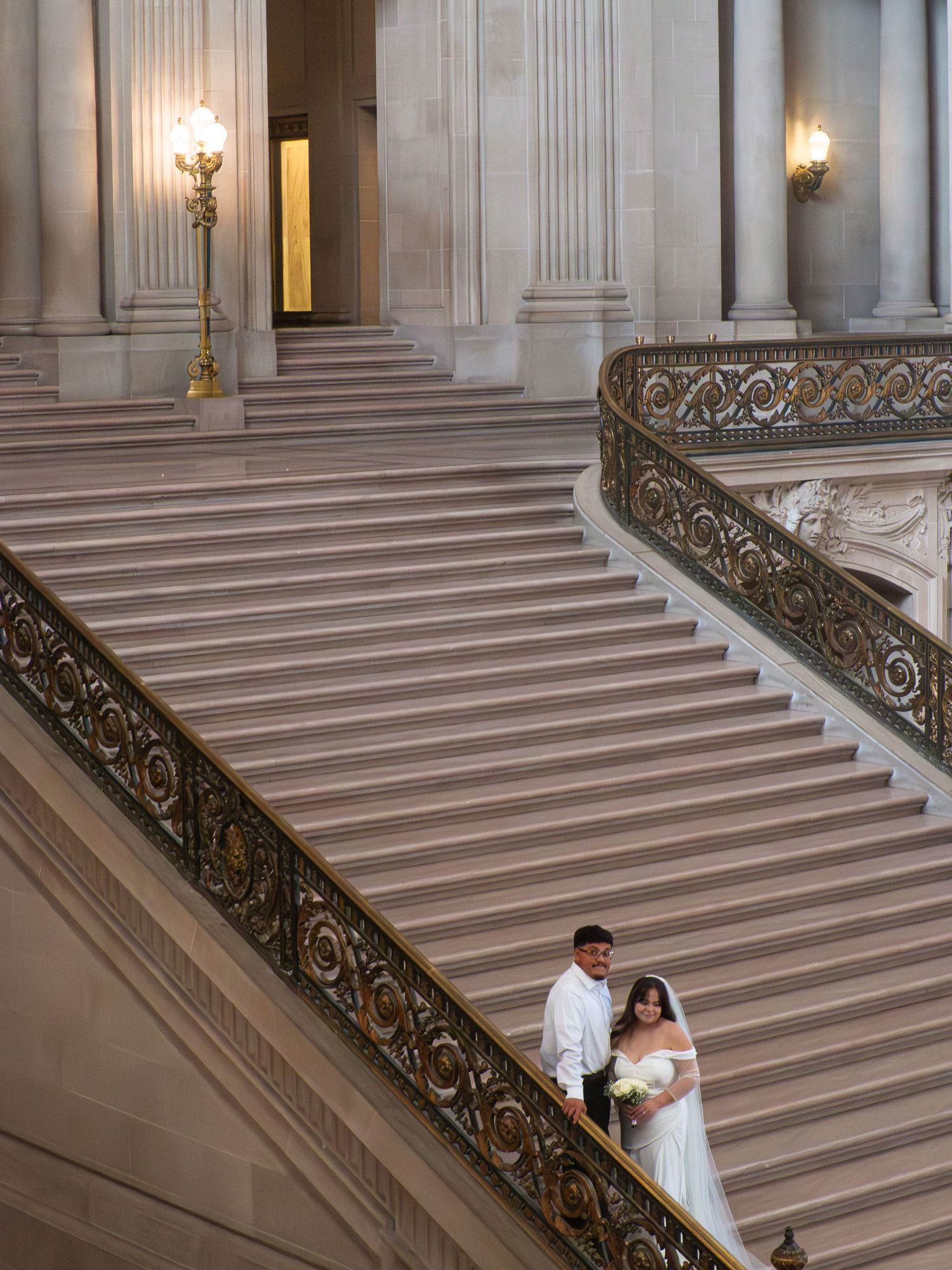 Newlywed couple poses on ornate grand staircase at San Francisco City Hall in wedding attire.