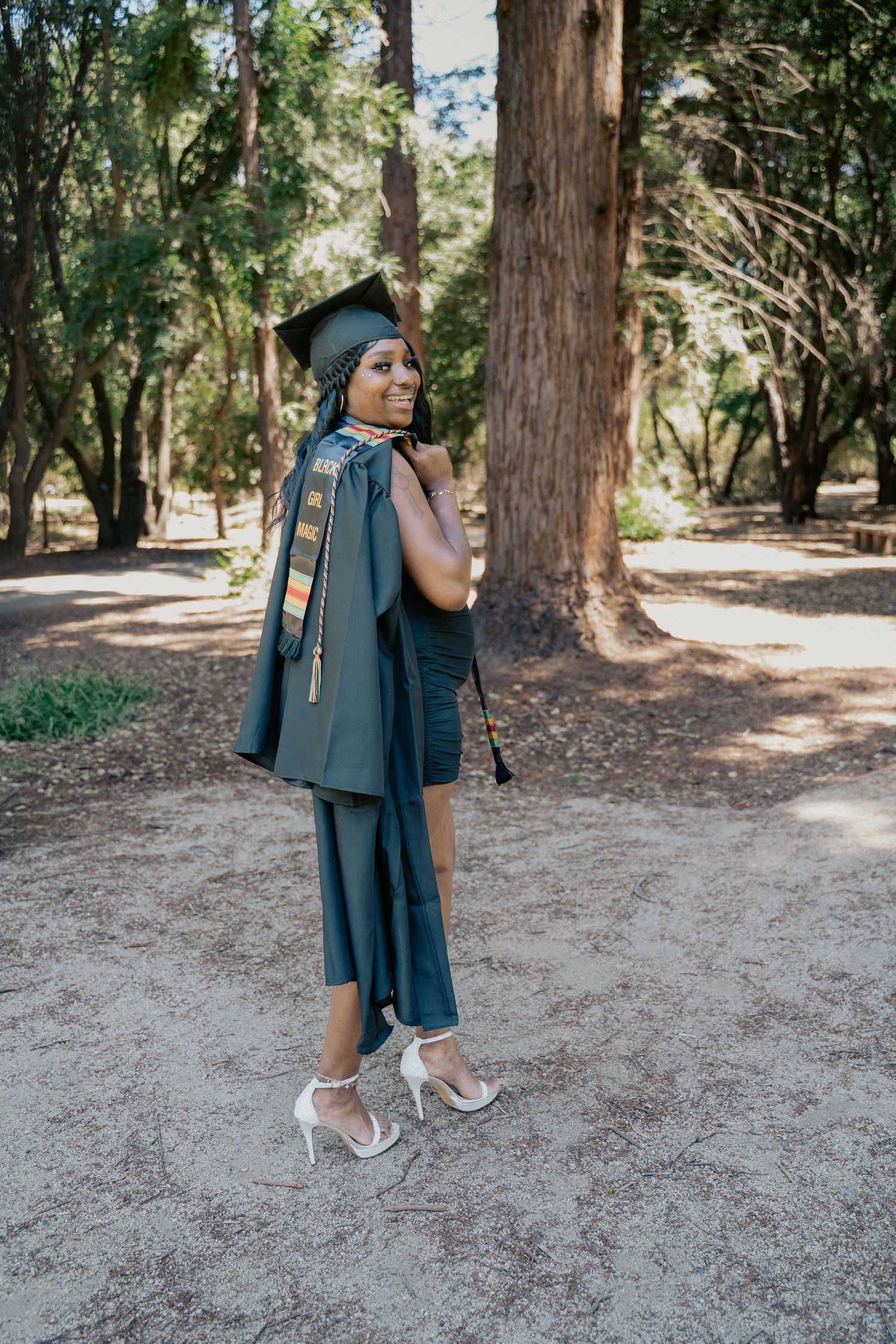 Young Black woman in green graduation cap and gown smiling over shoulder in wooded park setting.