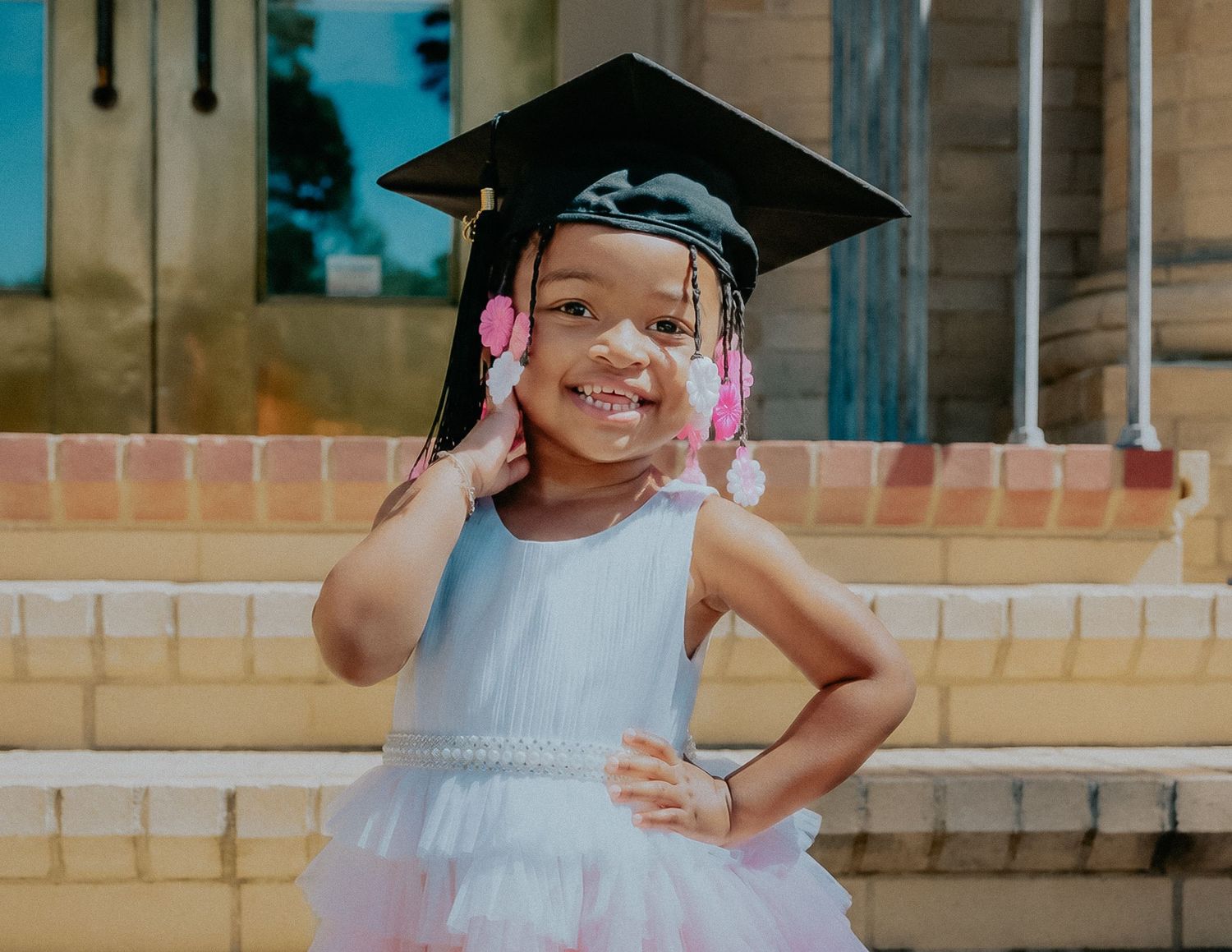 Adorable young girl in white tulle dress and graduation cap smiling with hand on hip outdoors.