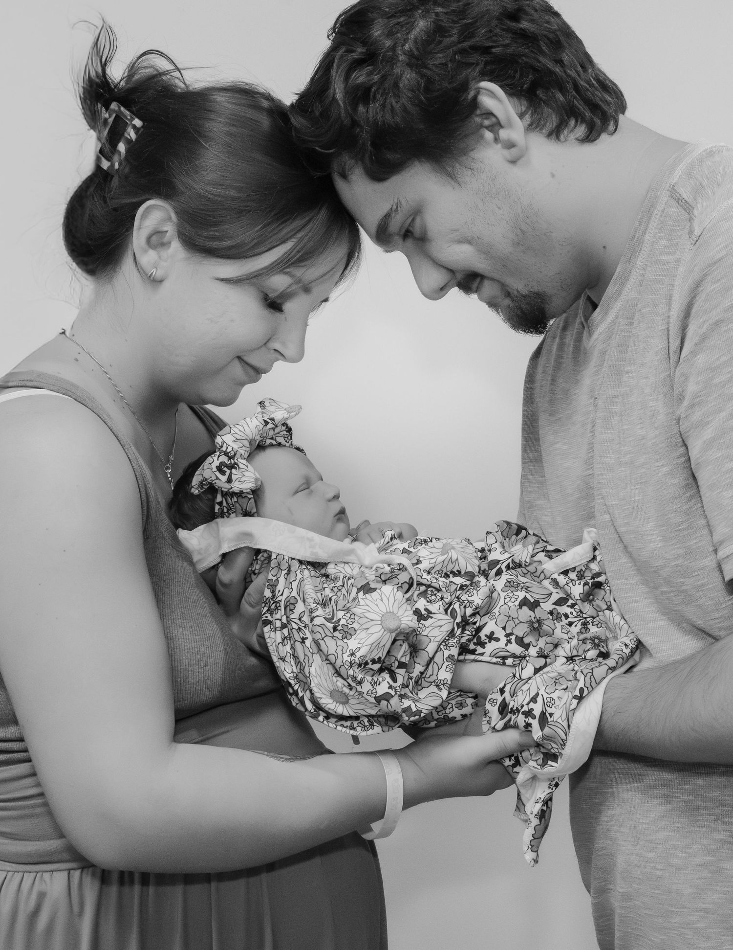 Black and white photo of new parents lovingly gazing at their newborn baby wrapped in a floral blanket.