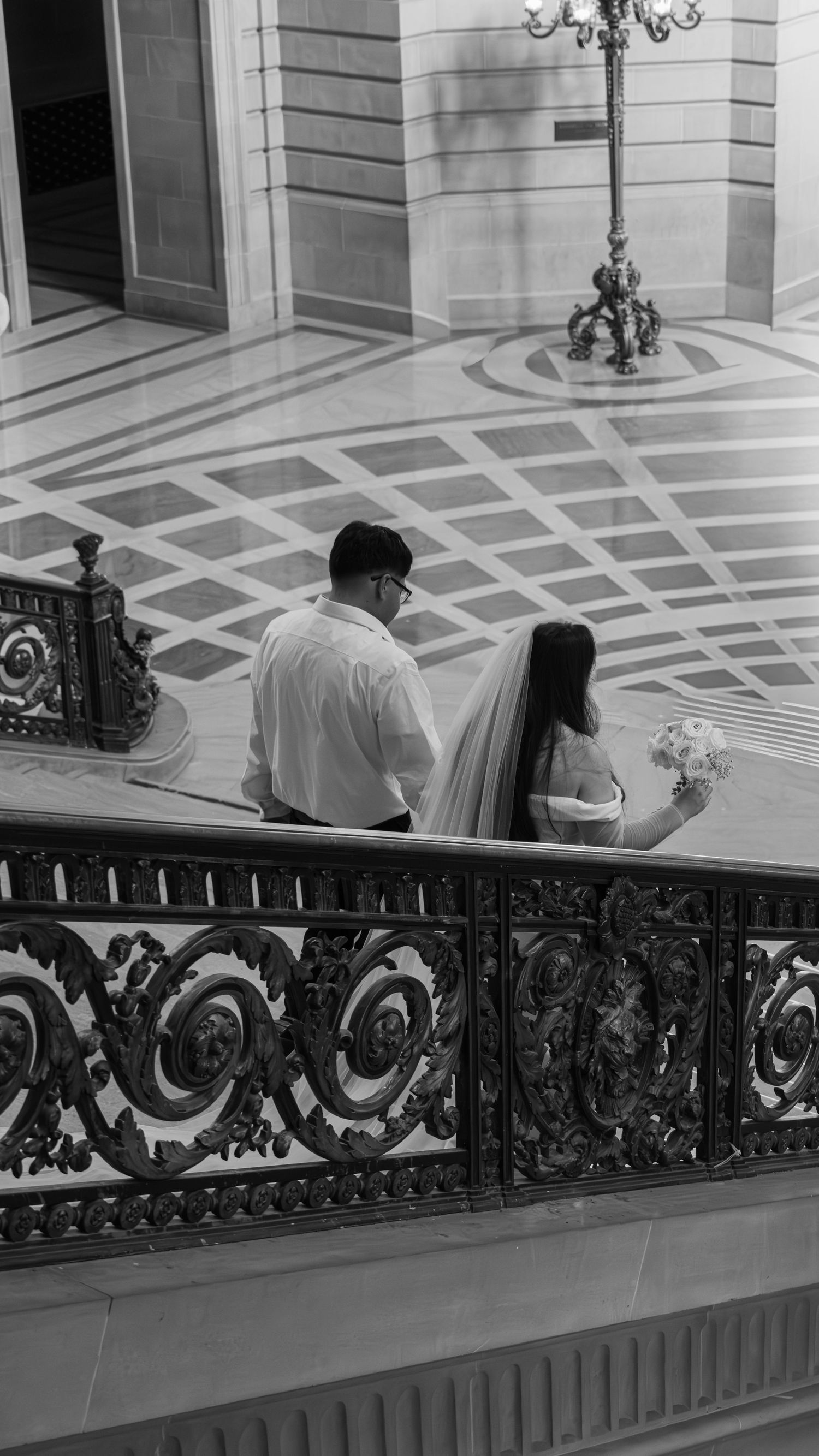 Black and white photo of a bride and groom walking through an ornate building with decorative iron railings. Elopemen photographer, Auburn,
