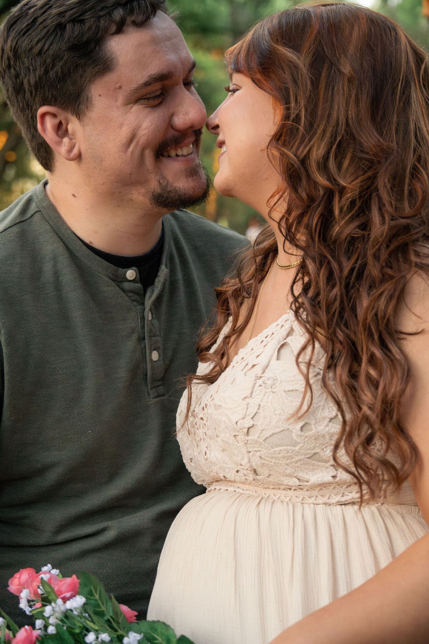 A romantic maternity photo shows a couple sharing a joyful moment outdoors while wearing green and cream colored attire.