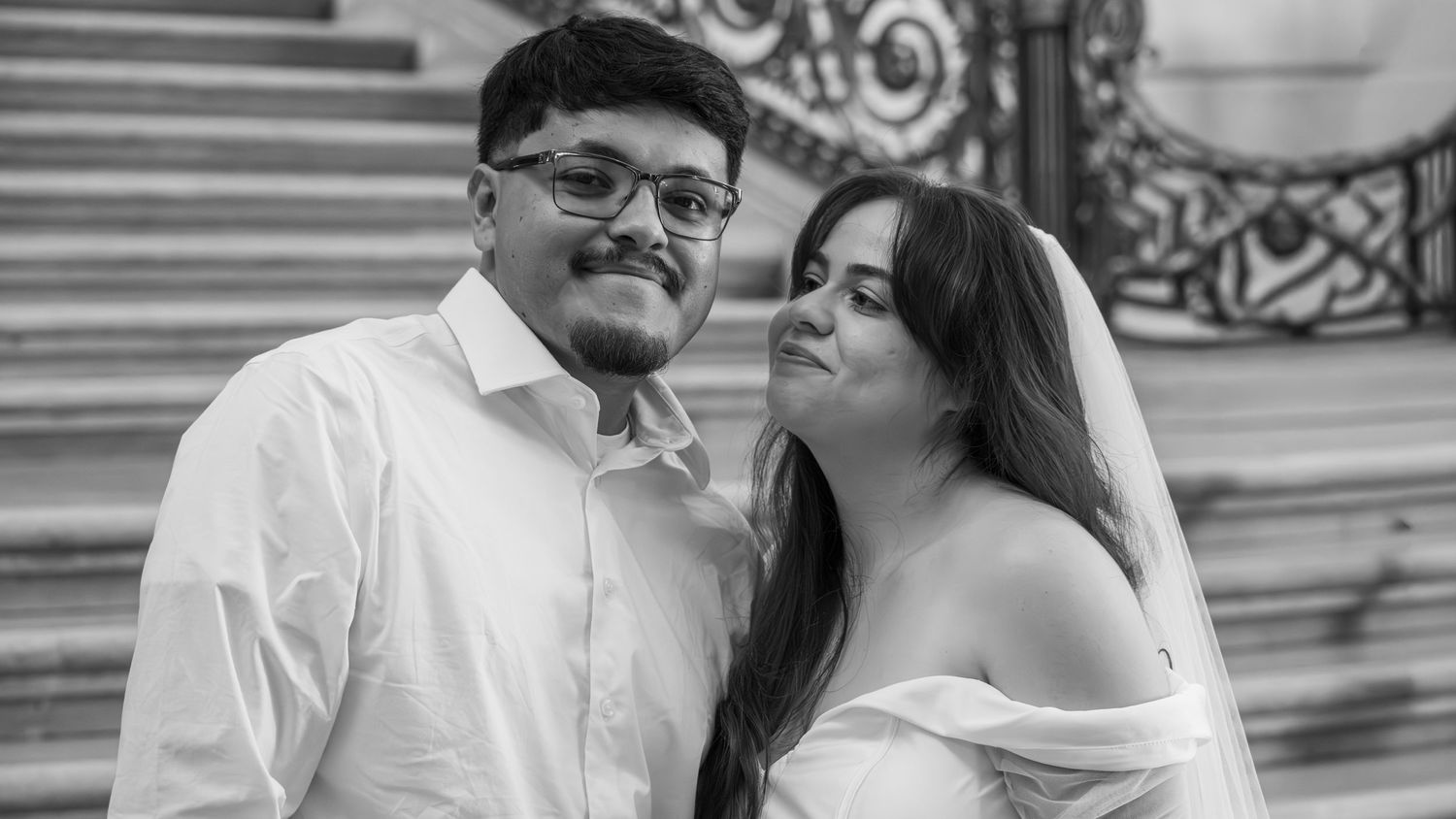 Black and white wedding photo of smiling couple, groom in white shirt and bride in off-shoulder dress on steps.