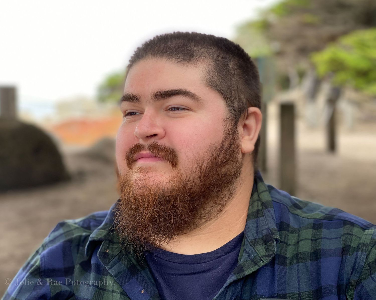 Portrait of a bearded man in a plaid flannel shirt smiling outdoors.