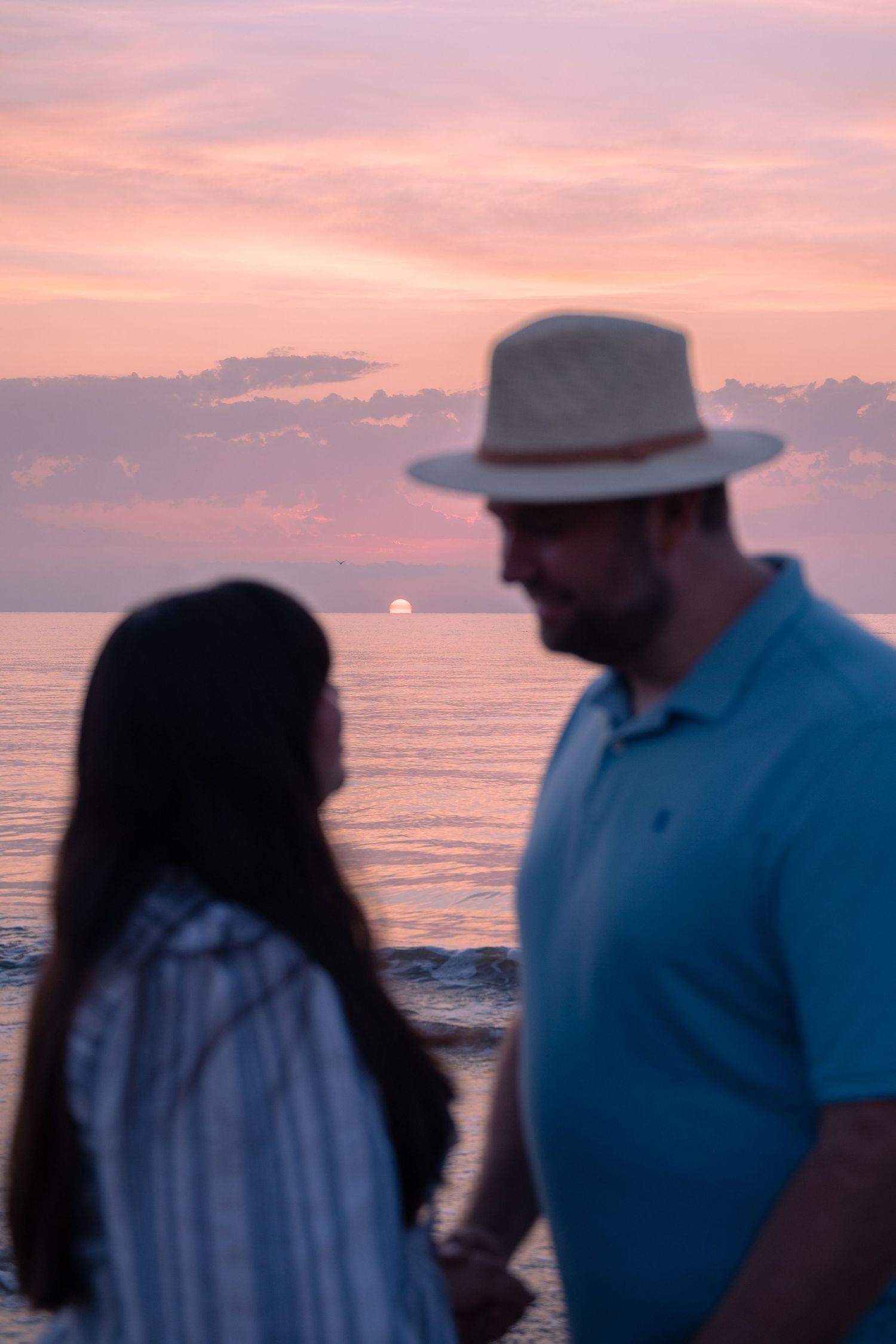 Two people walk along the shoreline at sunset with pink and orange sky.