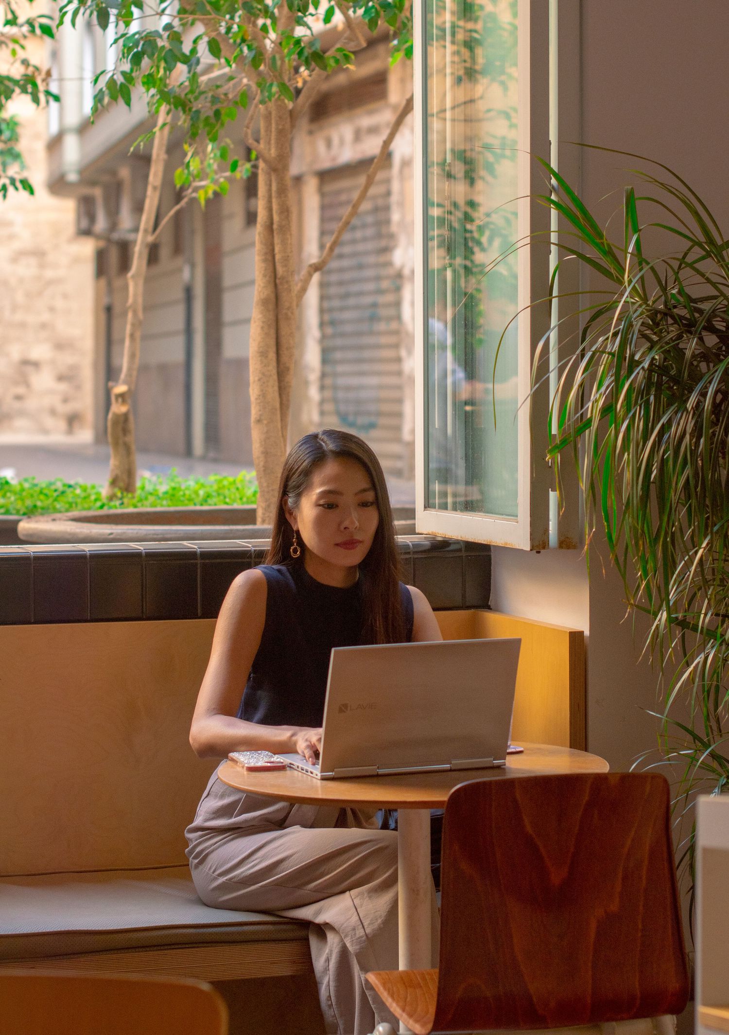 Person working on laptop in a cozy cafe corner with natural lighting and indoor plants.