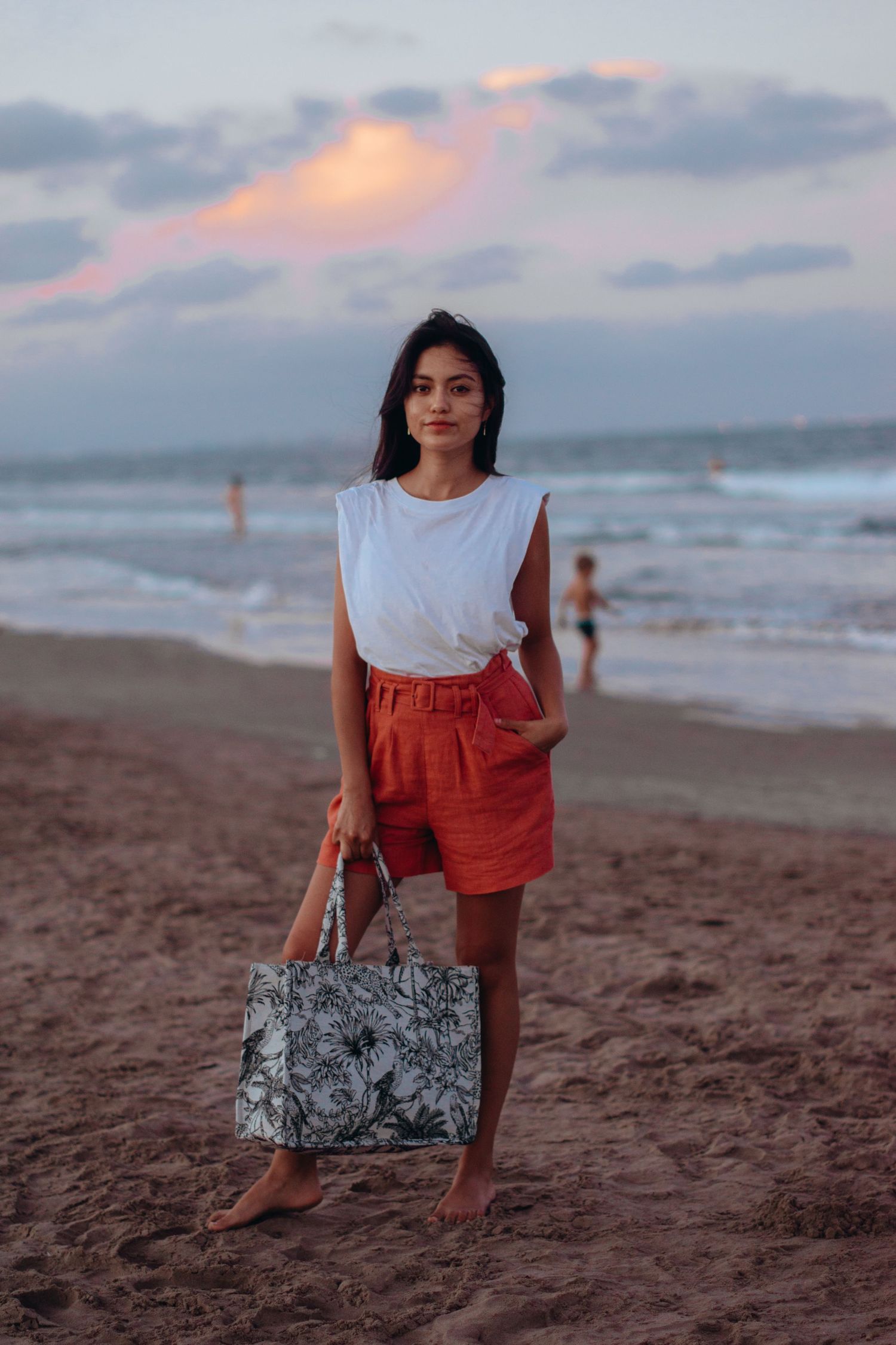 A person in summer attire sits on sandy beach with patterned bag at dusk.