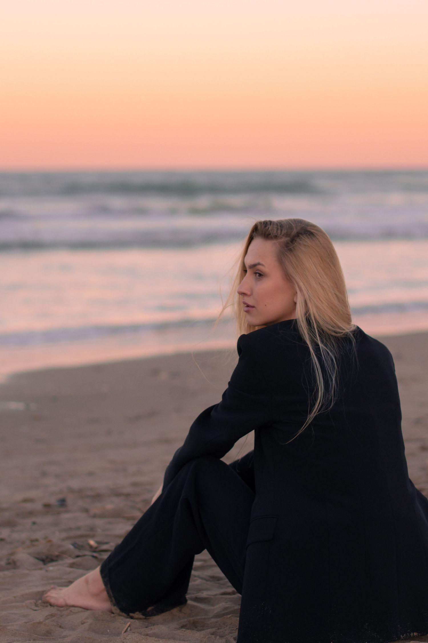 A figure in black clothing sits on beach sand during golden hour.