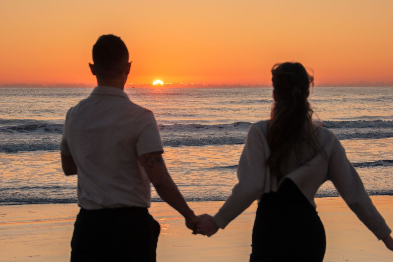 A couple walks hand in hand along the beach at sunset with golden light reflecting off the ocean waves.