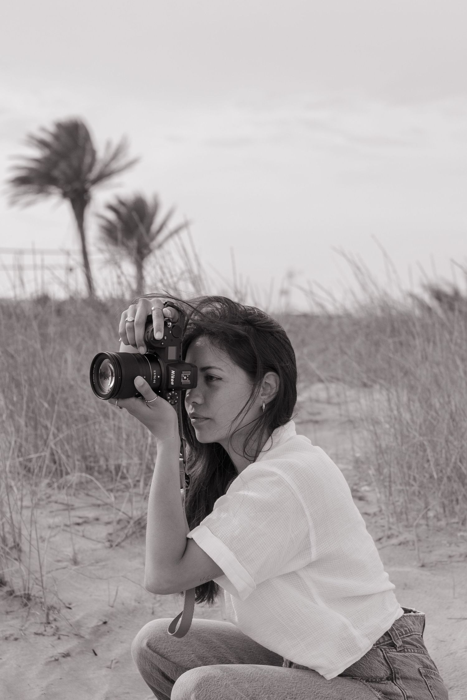 A photographer in a white t-shirt crouches on a beach with palm trees in the background, taking pictures in black and white.