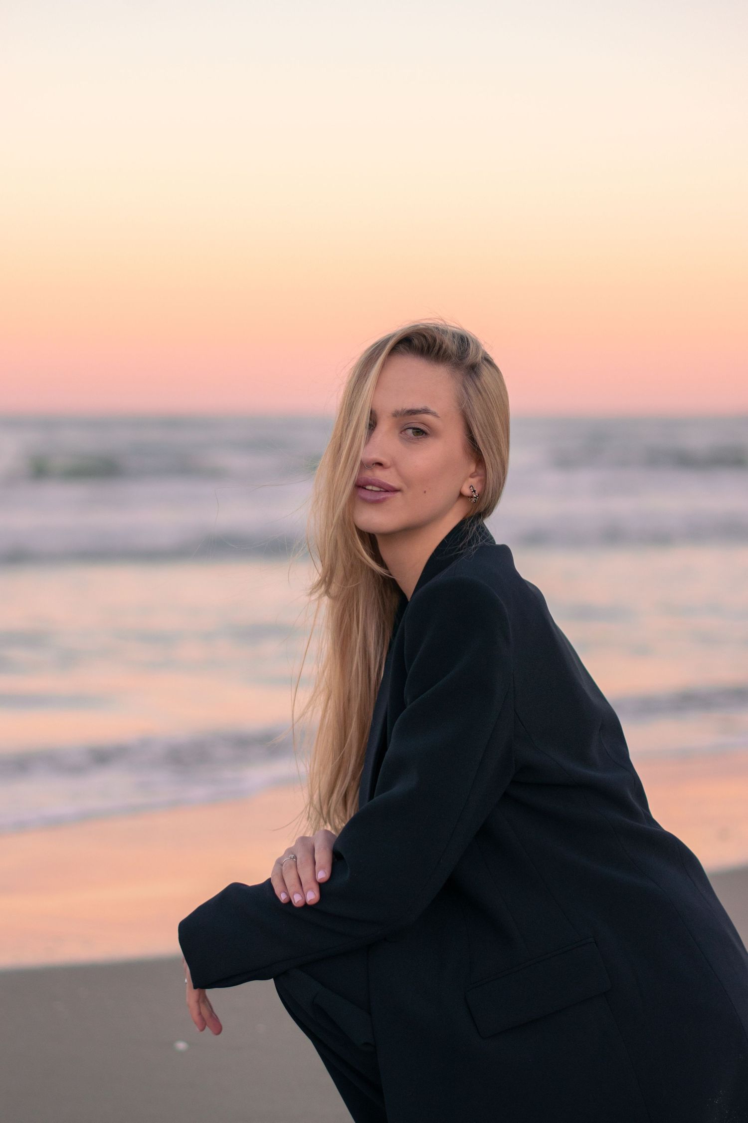 Blonde woman in black blazer poses on beach during golden sunset with ocean waves in background.