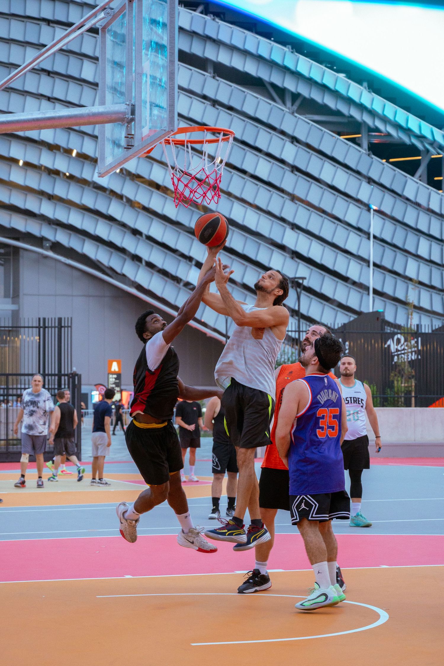 Basketball players compete for a shot at an outdoor court with a curved stadium roof in the background.