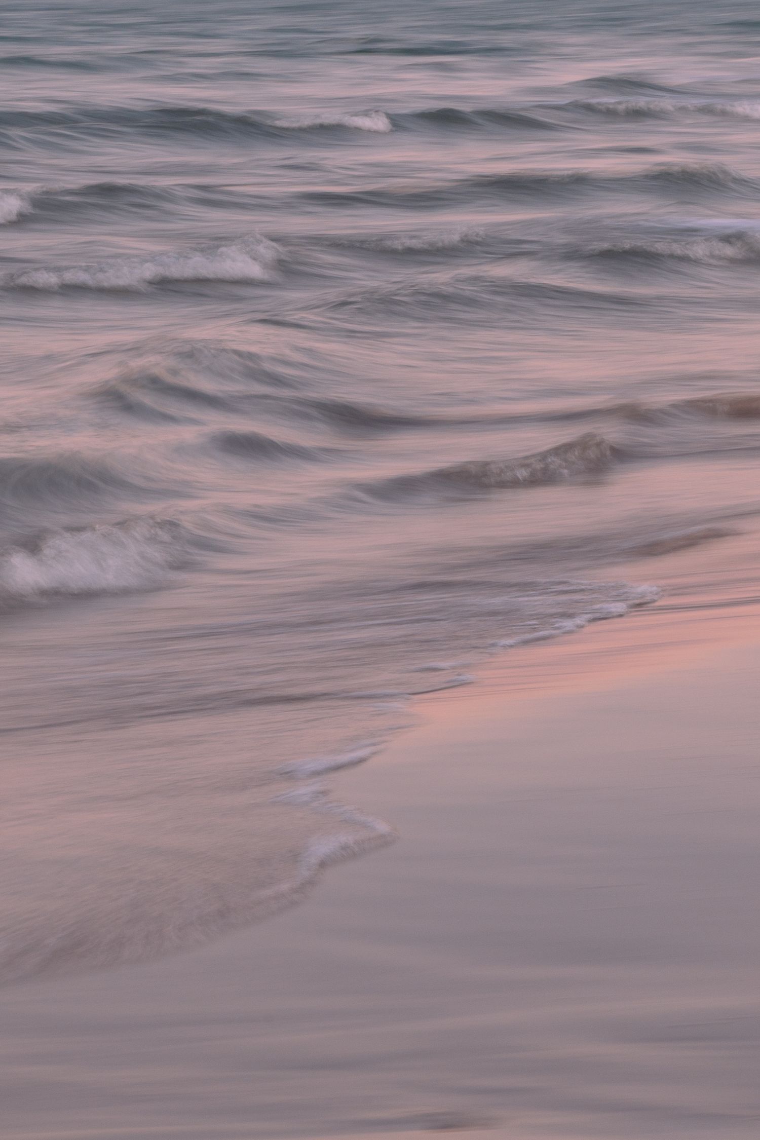 Gentle ocean waves ripple across the water's surface in soft pink and purple hues during sunset.