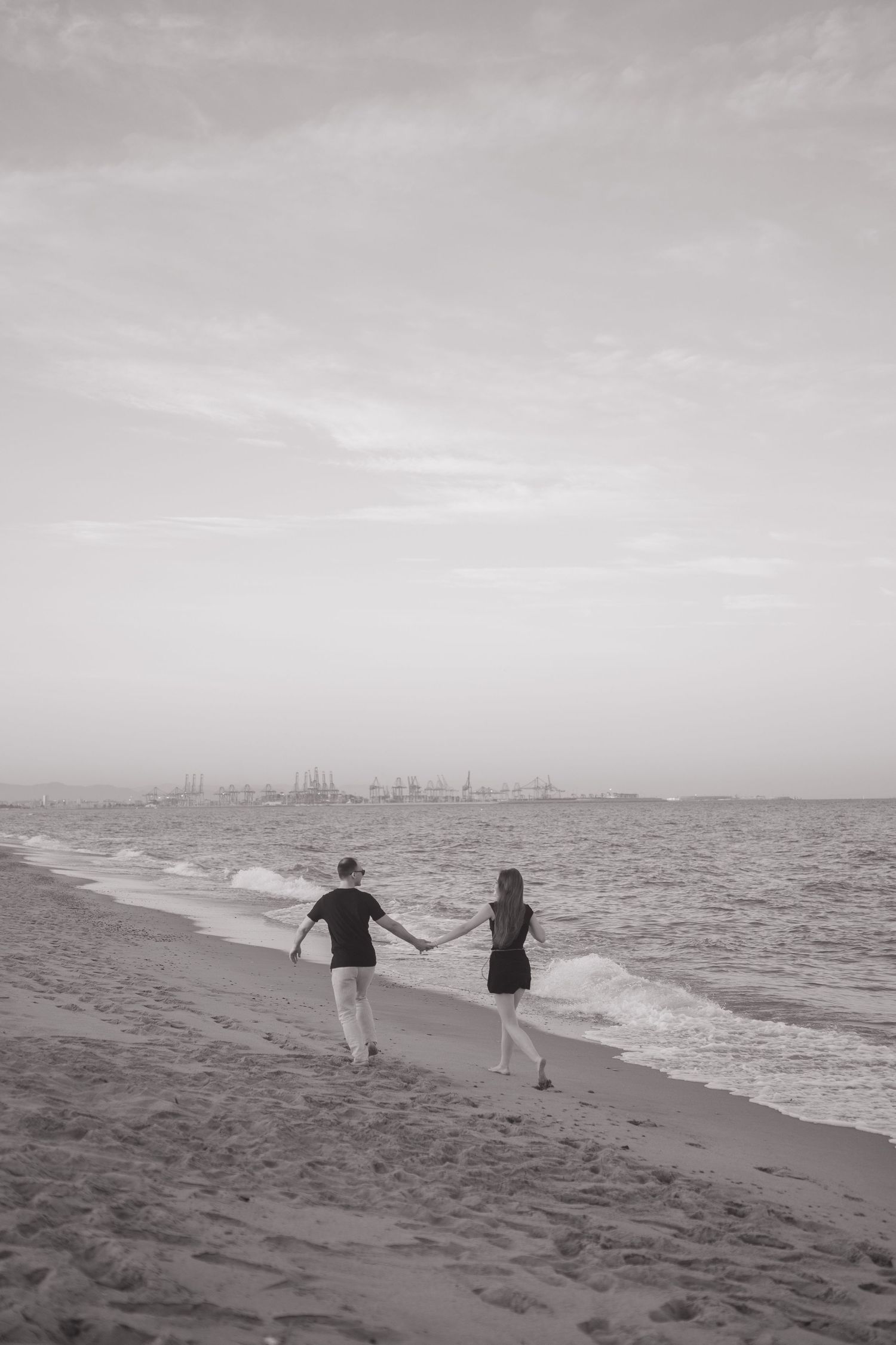 Two people walking hand in hand along a misty beach with a distant cityscape on the horizon.