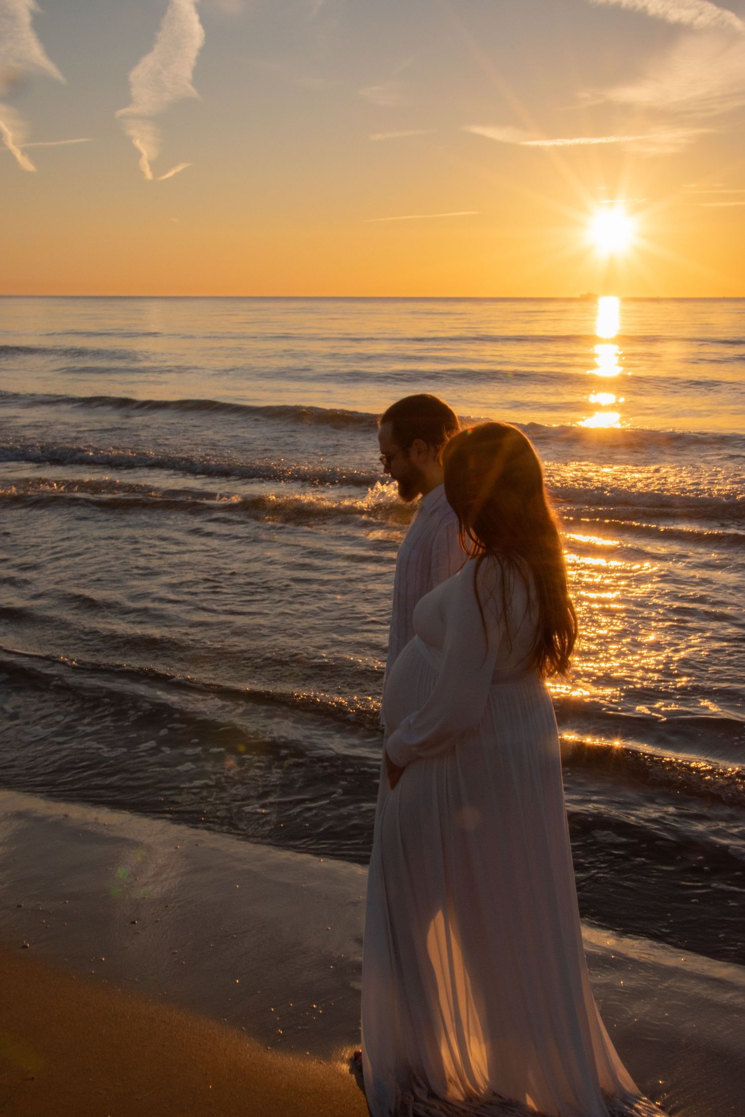 A couple in white dresses embraces on a beach at sunset with golden light reflecting off the ocean waves.