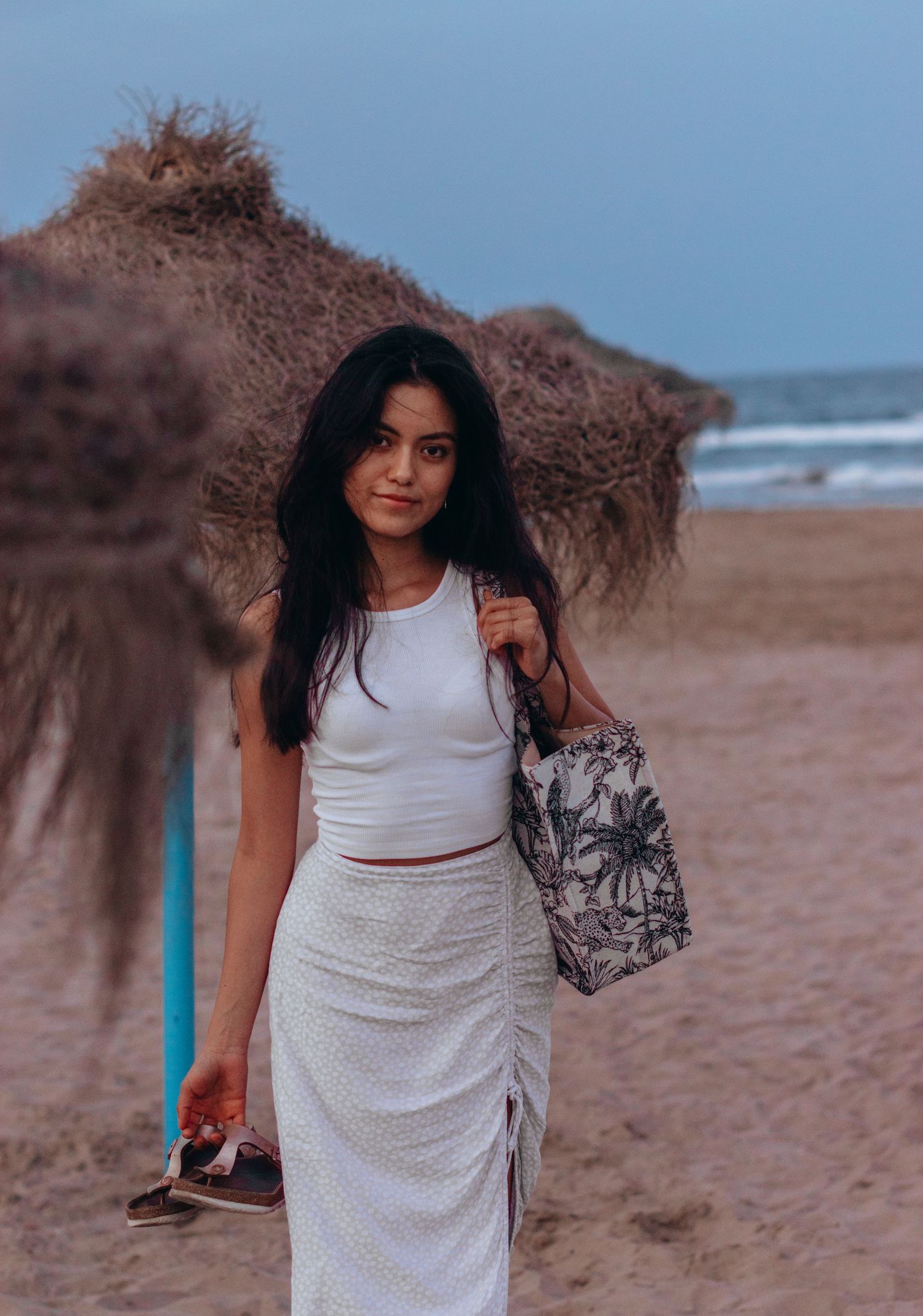 Woman in white dress walks along beach with curly hair blowing in wind.