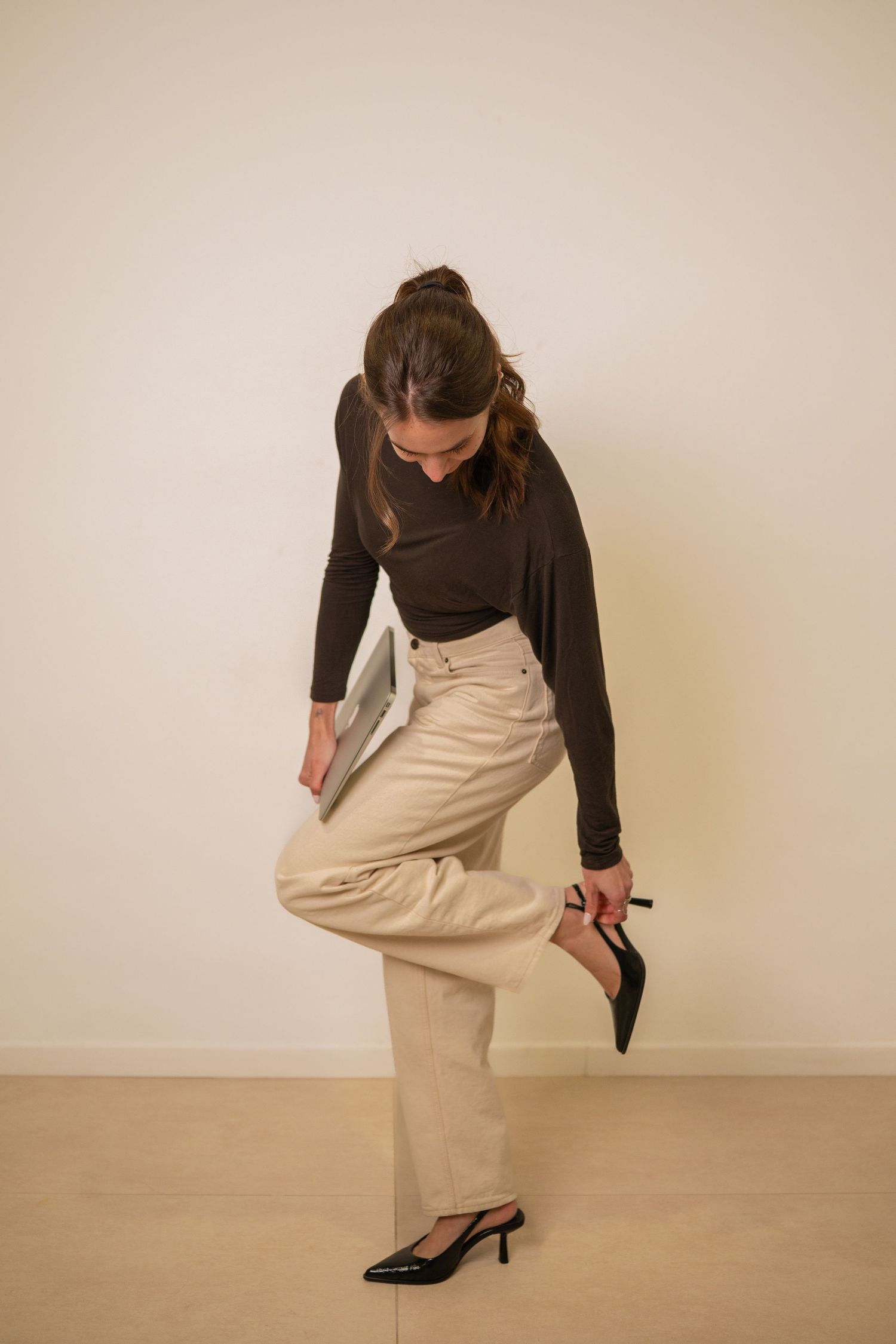 Woman in black top and beige skirt adjusting shoe in minimalist setting.